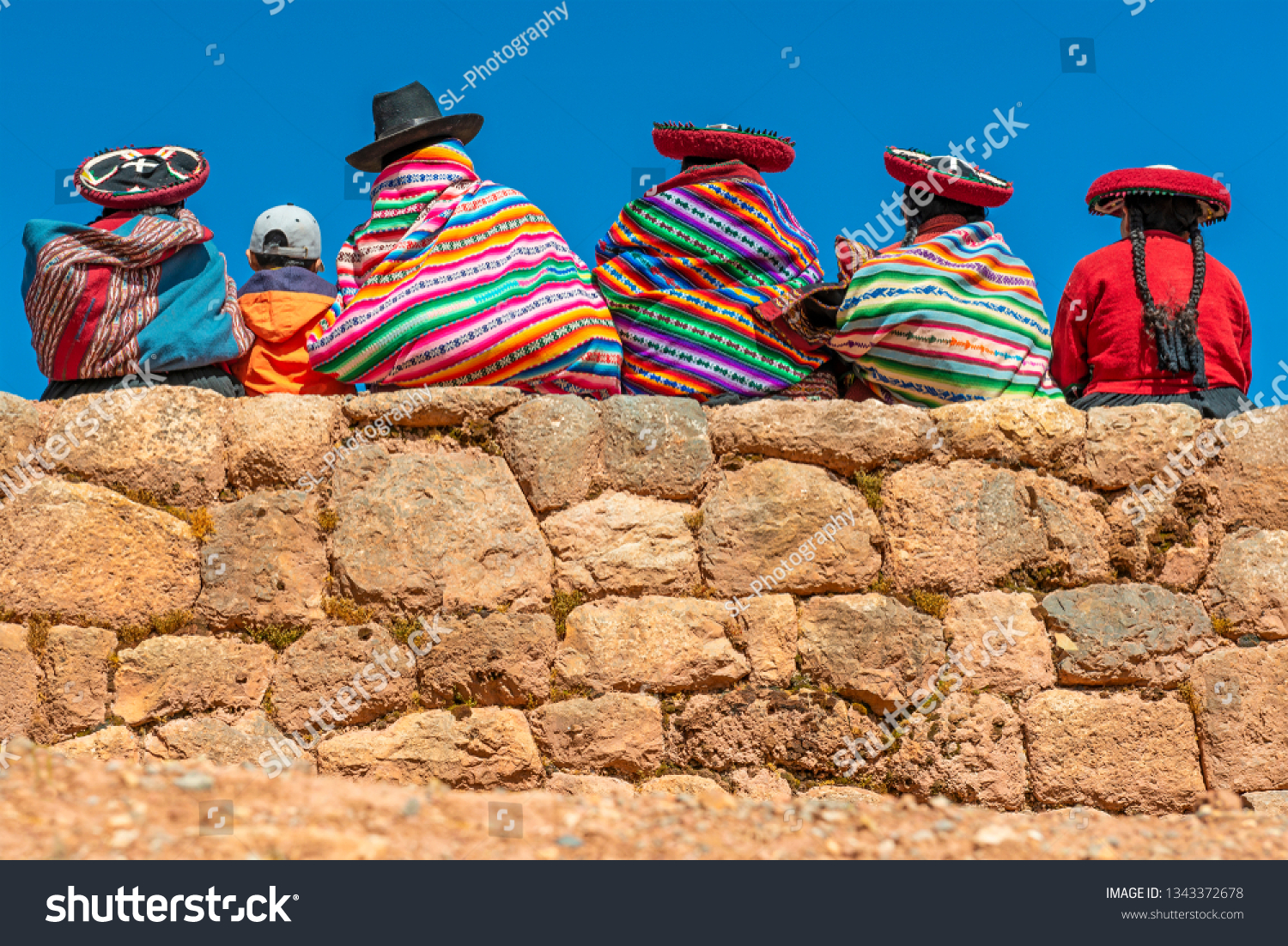 A group of Quechua indigenous women in traditional clothing and a young boy sitting and chatting on an ancient Inca wall in the archaeological site of Chinchero in the region of Cusco city, Peru. #1343372678