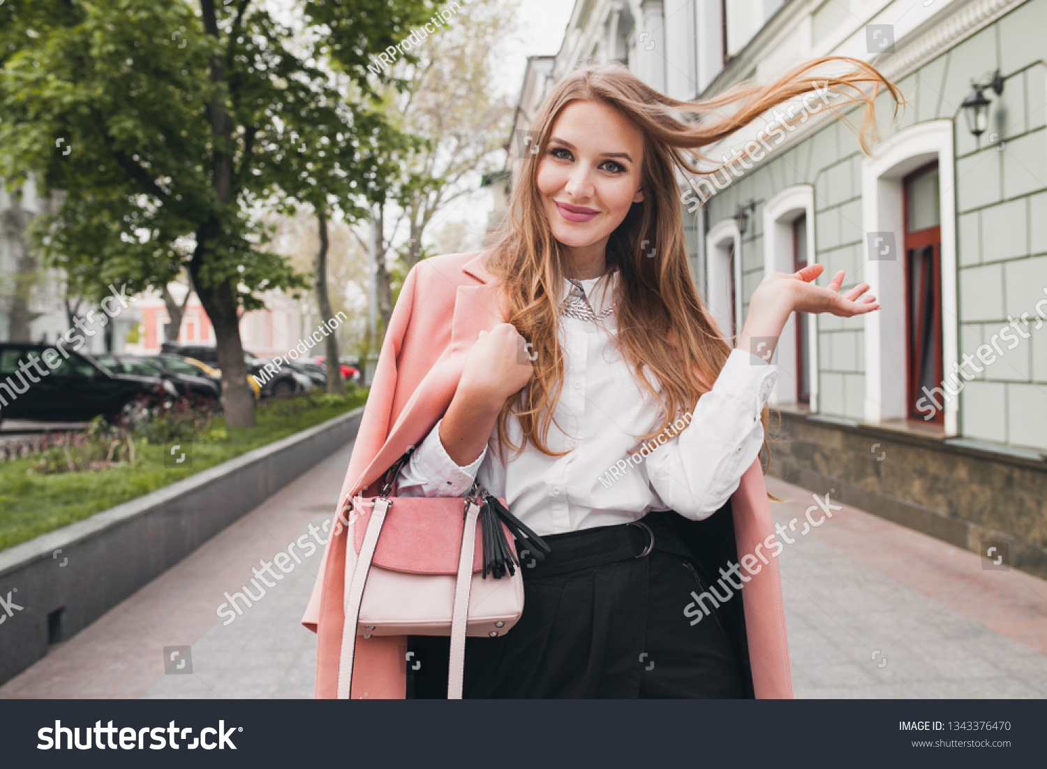 attractive stylish smiling woman walking city street in pink coat spring fashion trend holding purse, elegant style, waving long hair #1343376470