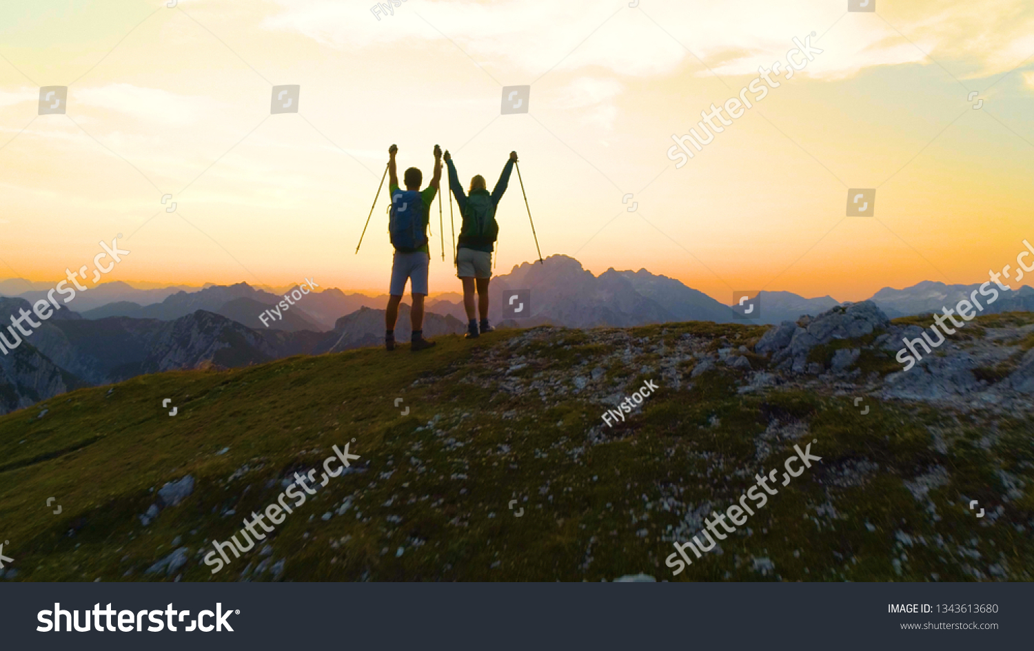 AERIAL: Unrecognizable young hiker couple outstretch arms as they catch the sunset while hiking in the Julian Alps. Picturesque shot of golden sunrise illuminating the excited male and female trekkers #1343613680