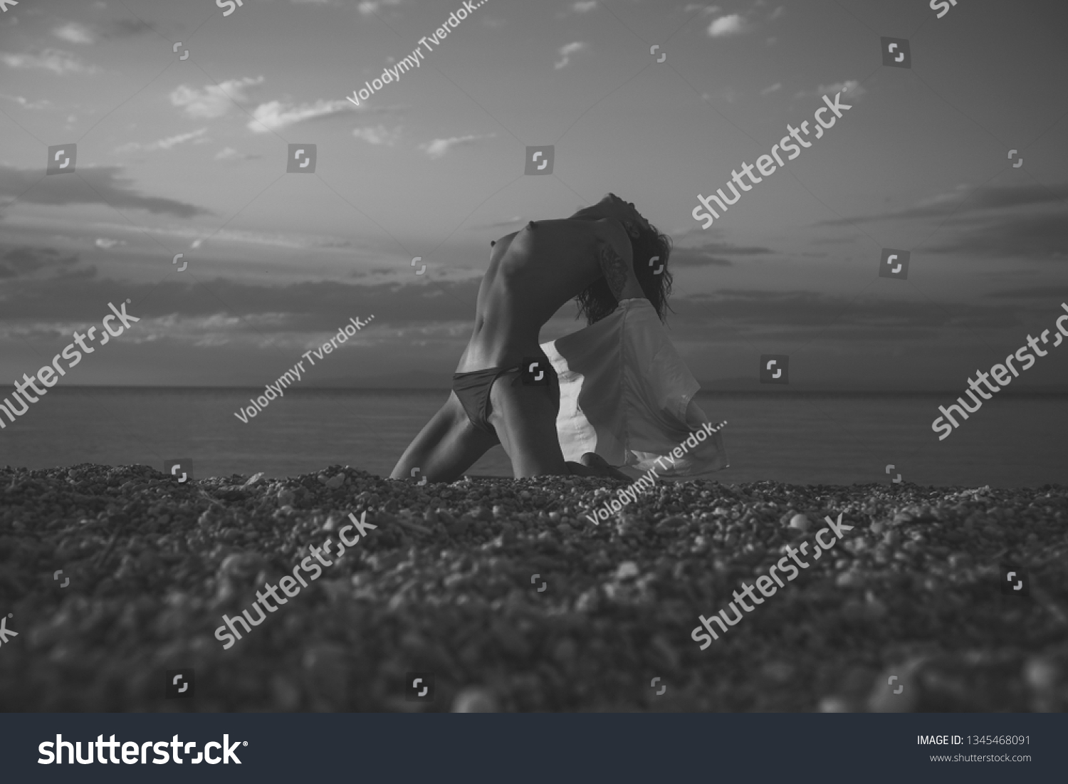Attractive young lady suntanning nude, nudist, enjoy last sun rays. Girl sexy, topless, naked breasts with wild hair at seashore at sunset. Erotic concept. Woman stands on knees on beach in evening. #1345468091