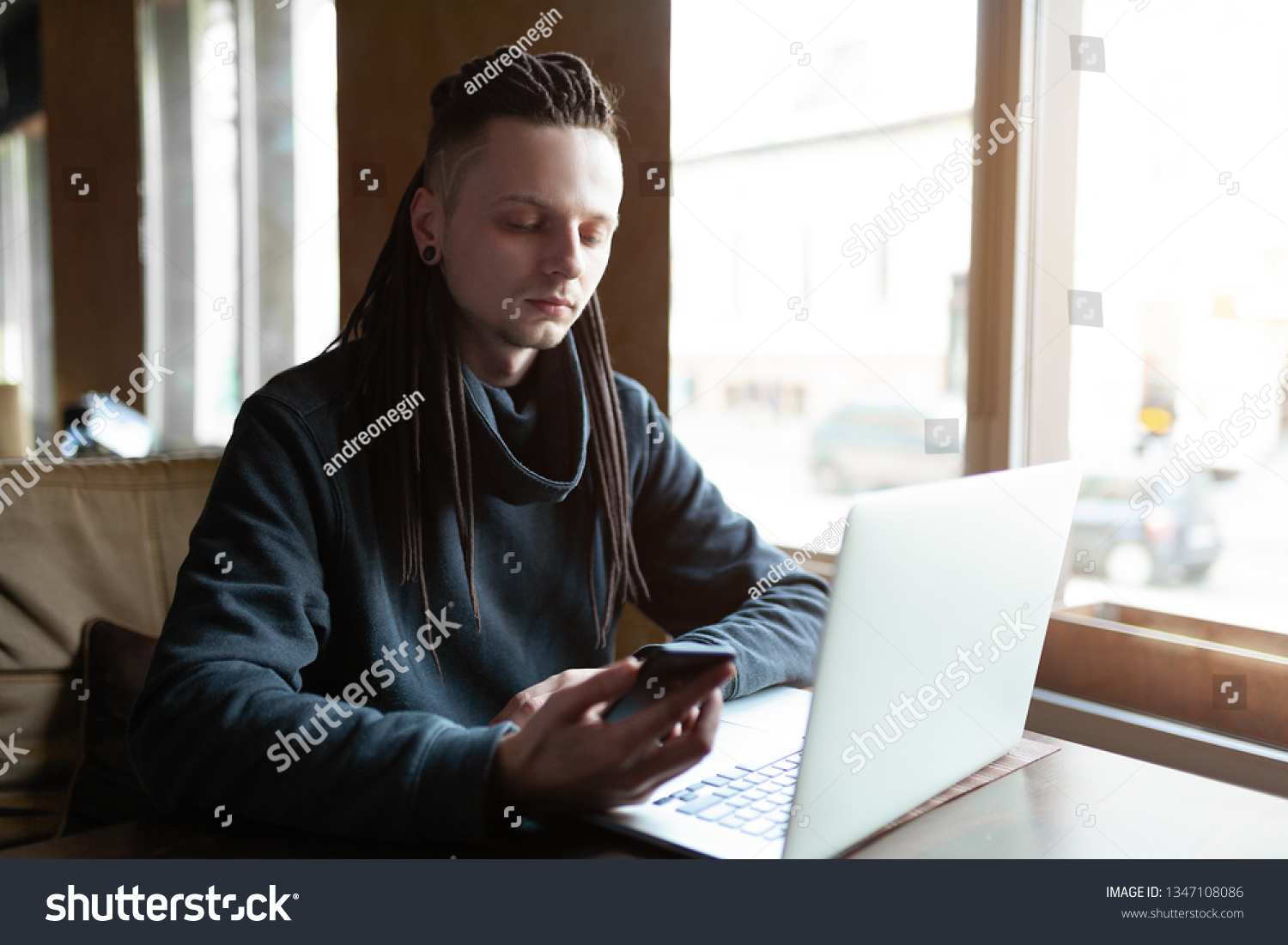 Young Businessman with dreadlock having doing his work in cafe near the big window with laptop. Youth culture. Neformal guy. Remote works. IT specialist #1347108086
