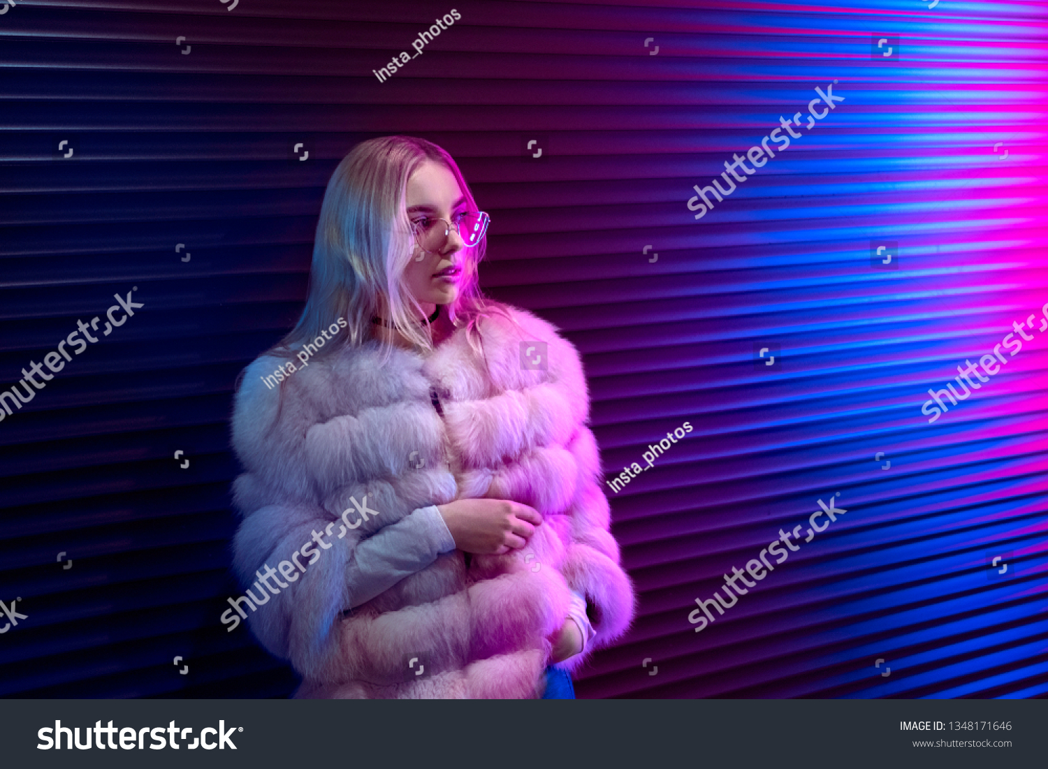 Teen hipster girl in stylish glasses and fur standing on purple street neon light wall background, female teenager fashion model pretty young woman looking at night club city light glow, back to 80s #1348171646