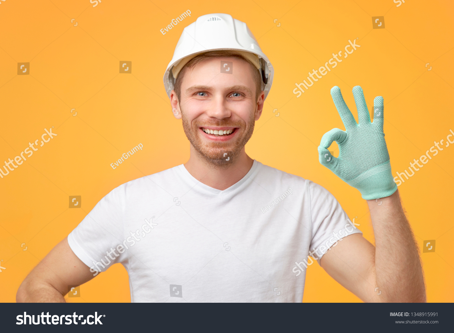 Pleased confident European man with broad smile, uniform, shows okay gesture, dressed in t-shirt and construction helmet. Isolated over white background.  #1348915991