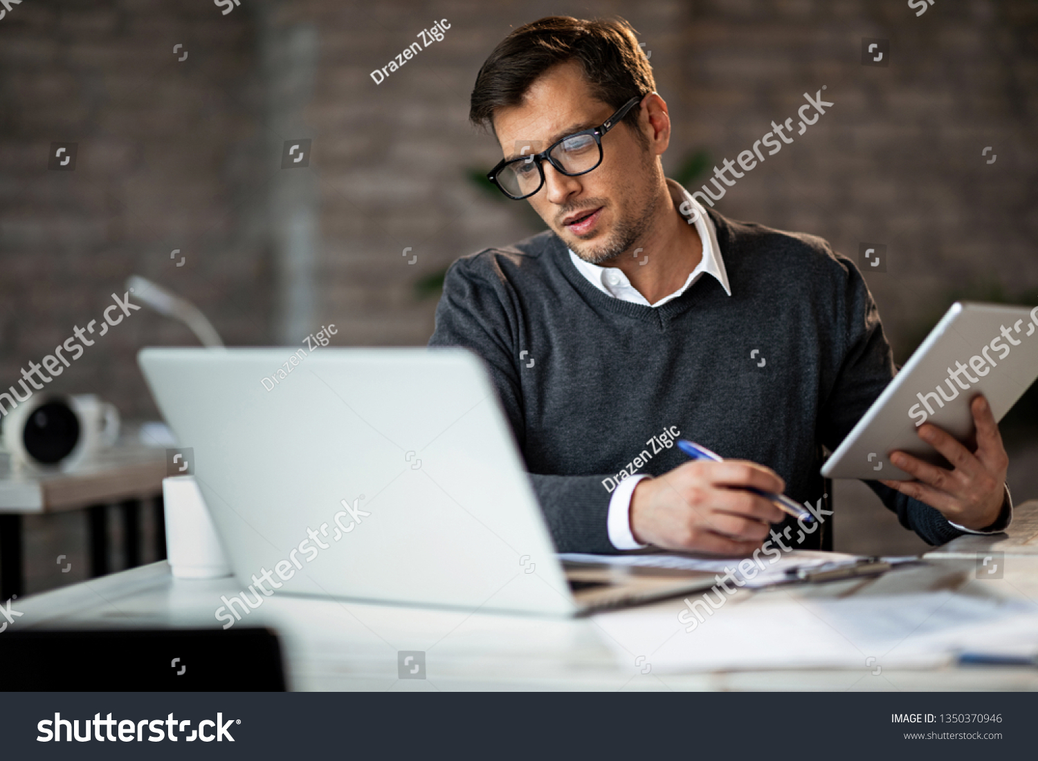 Multi-tasking businessman working in the office. He is using touchpad while reading an e-mail on laptop and taking notes on the paper. #1350370946