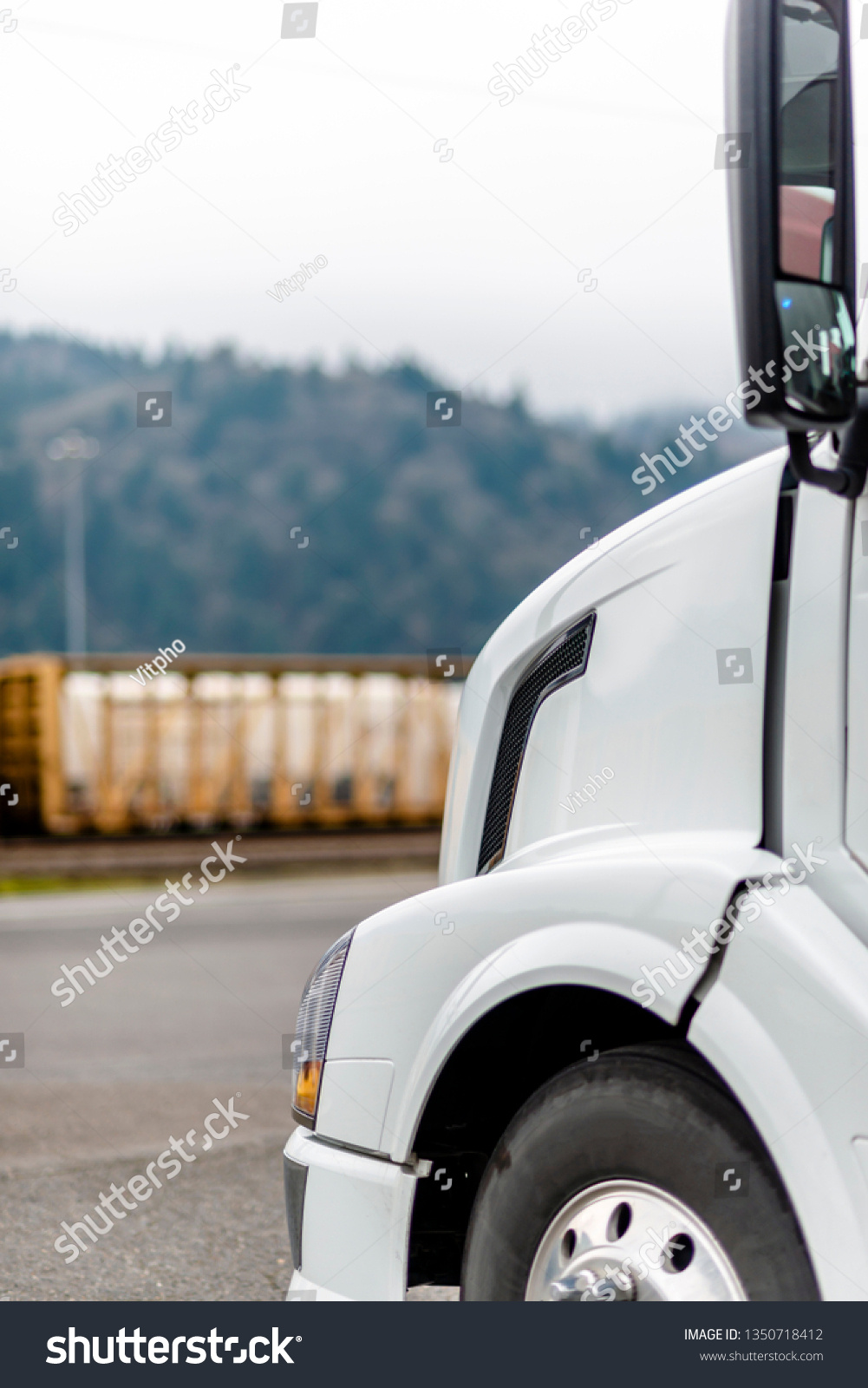 The front of professional industrial grade cab and hood of modern big rig white clean semi truck with front wheel parked on parking lot near the warehouse waiting commercial cargo load for delivery #1350718412
