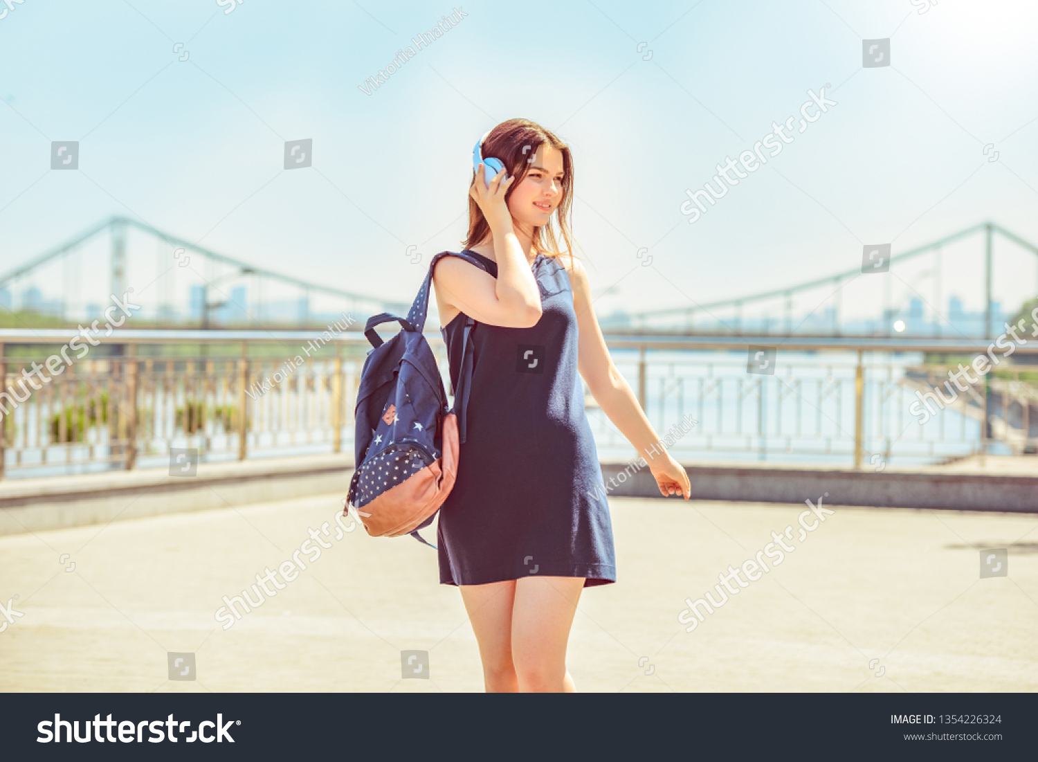 Teenager girl wearing headphones carrying backpack walking on the city street enjoying music smiling happy #1354226324