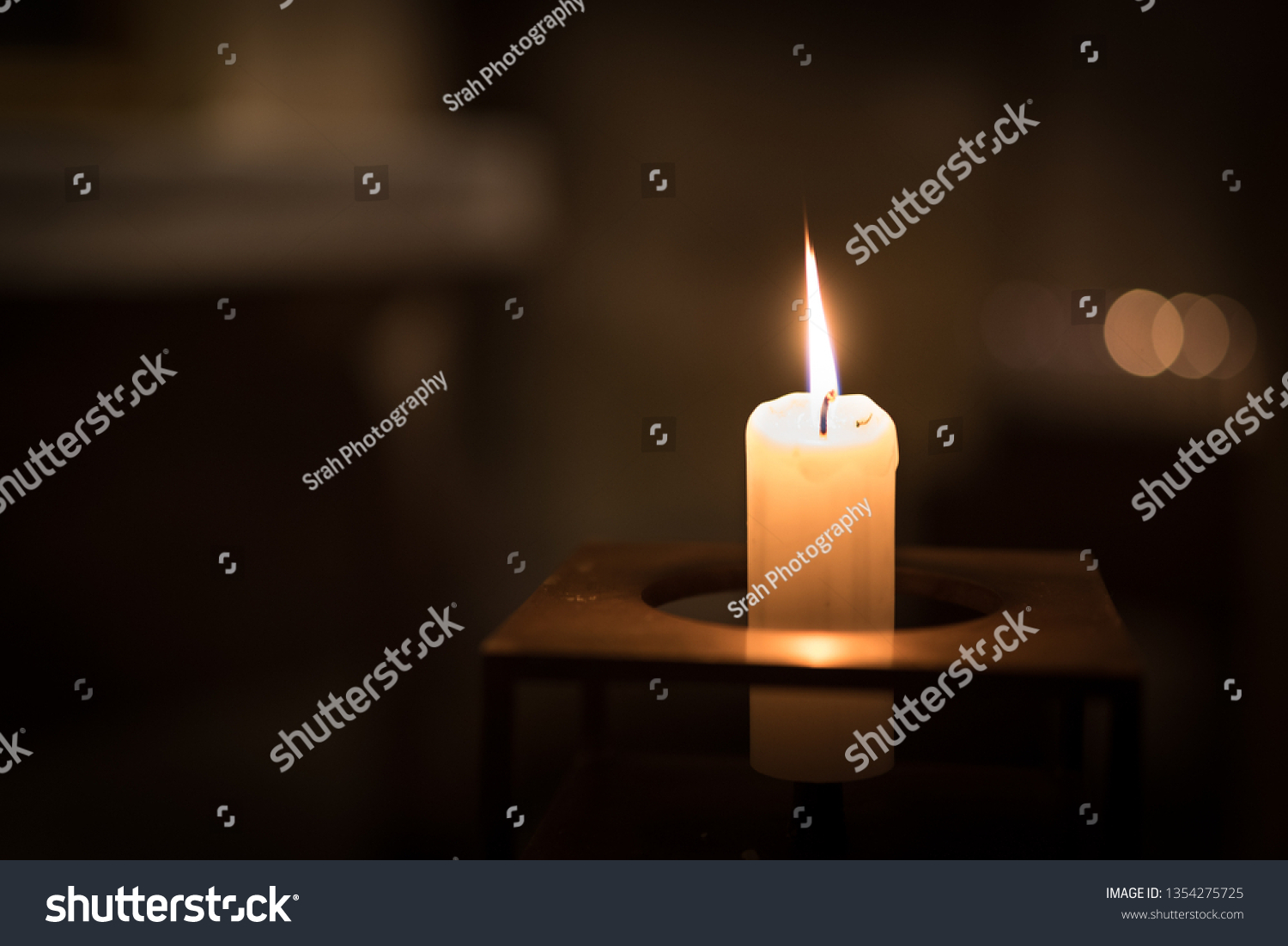 A single candle burning inside a church. The candle is made of pale wax and is in a square metal candle holder. The candle flame is sharp, and the background has soft bokeh. #1354275725