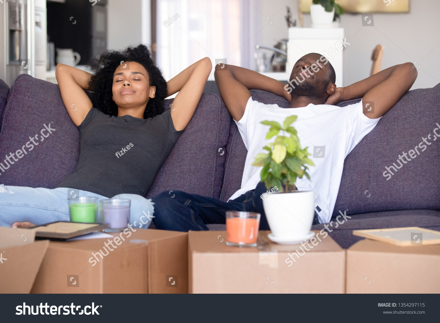 African couple put hands behind head rest on sofa with closed eyes big boxes with stuff, breath fresh air take break on moving day into new first home. Property buyers or easy delivery service concept #1354297115