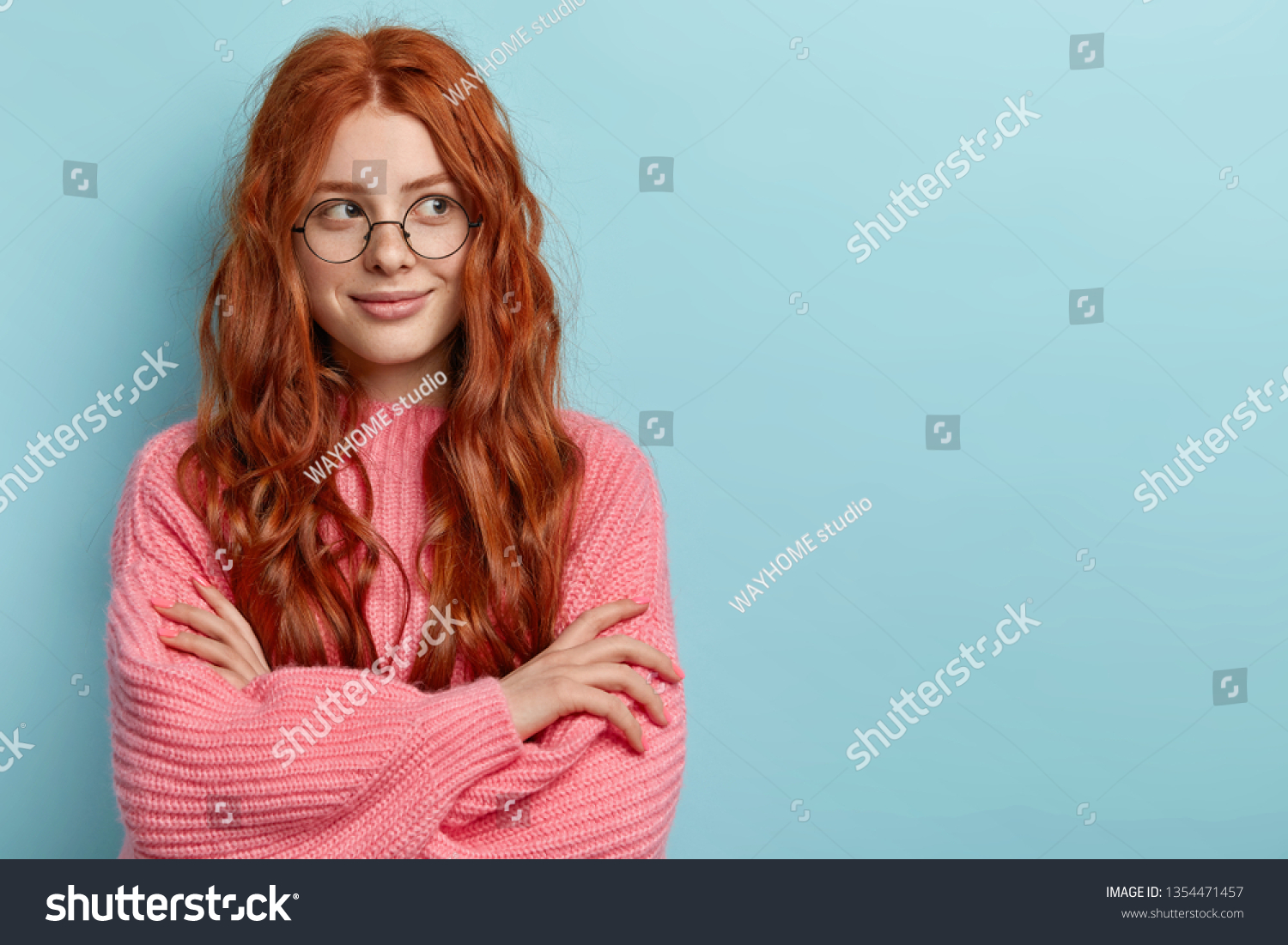 Pensive dreamy lovely young ginger female with pleasant appearance, imagines dreams come true, keeps hands crossed, dressed in oversized jumper, wears optical glasses, isolated over blue wall. #1354471457