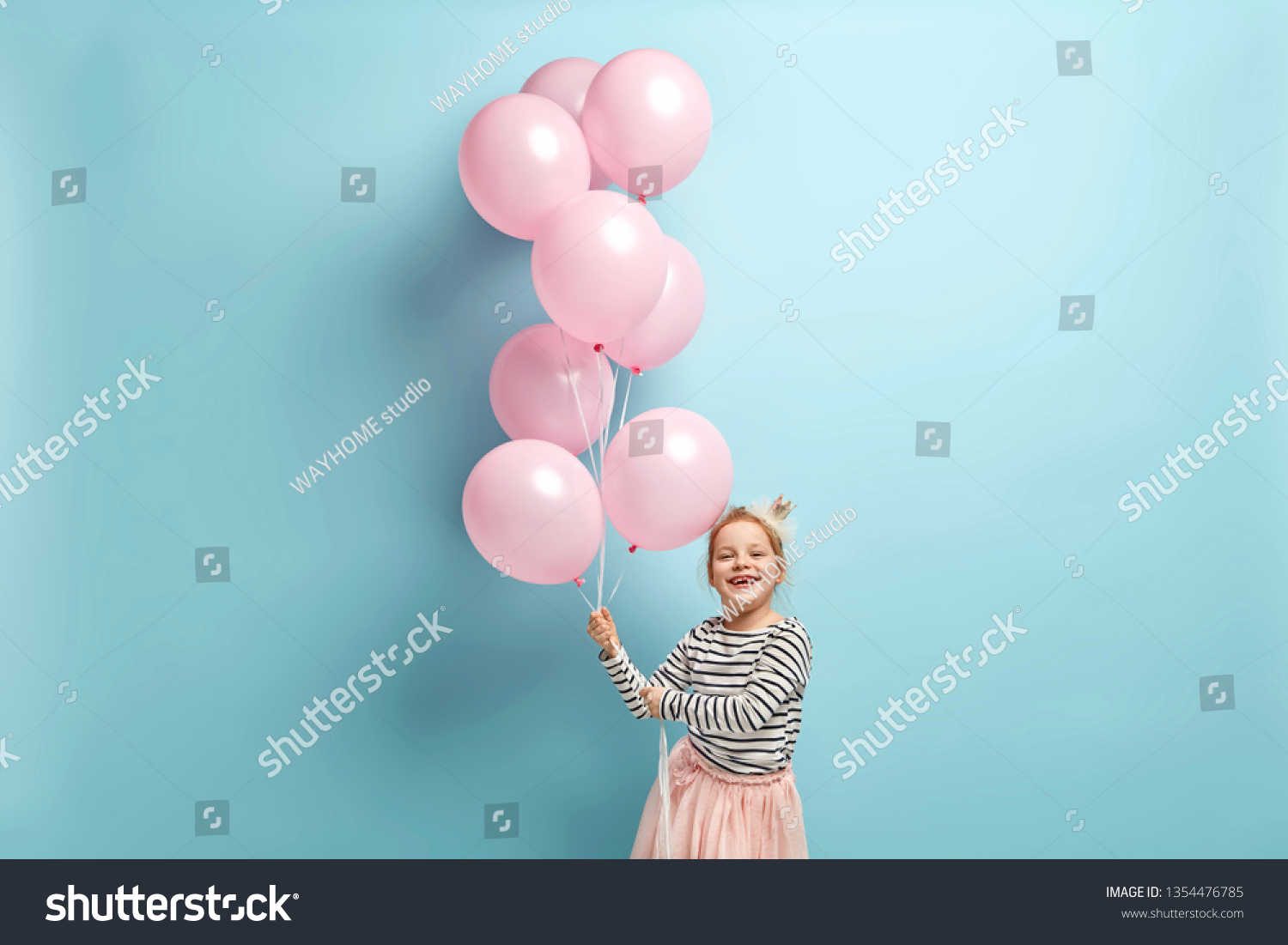Positive small kid dressed in festive clothes, carries air balloons, celebrates holiday, has broad smile, stands against blue background, being in high spirit. Children and celebration concept #1354476785