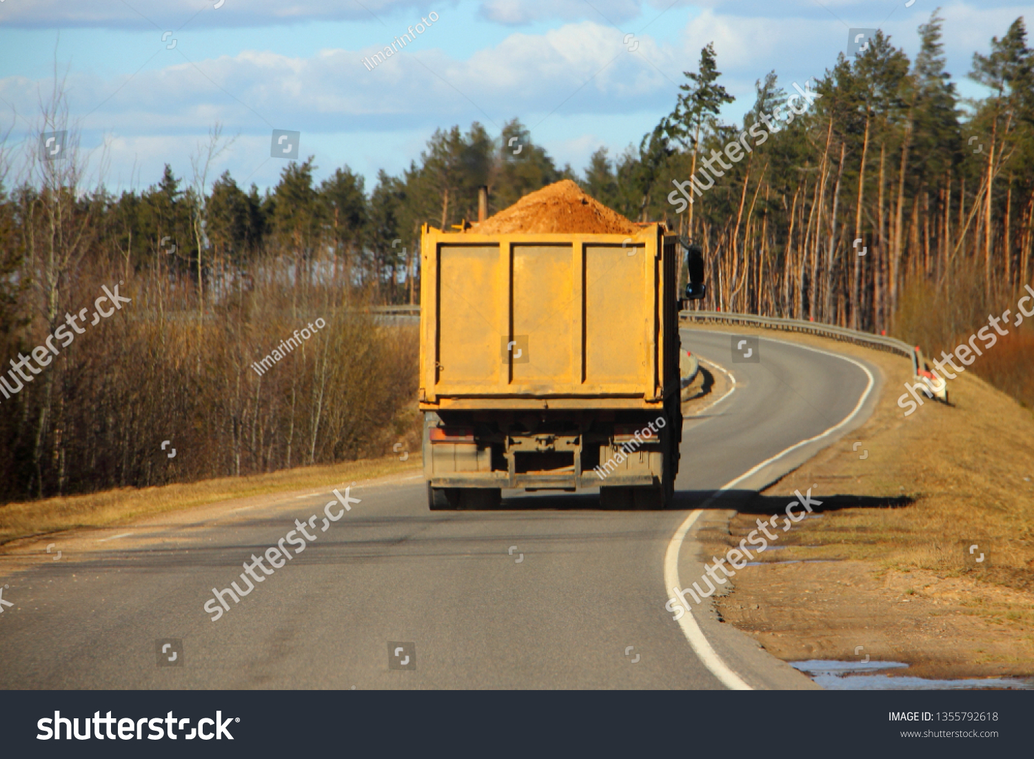 Transportation of bulk building materials, heavy overloaded dump truck carries sand on a European countryside one way asphalt serpentine road turn on pine forest and blue sky background at autumn day #1355792618
