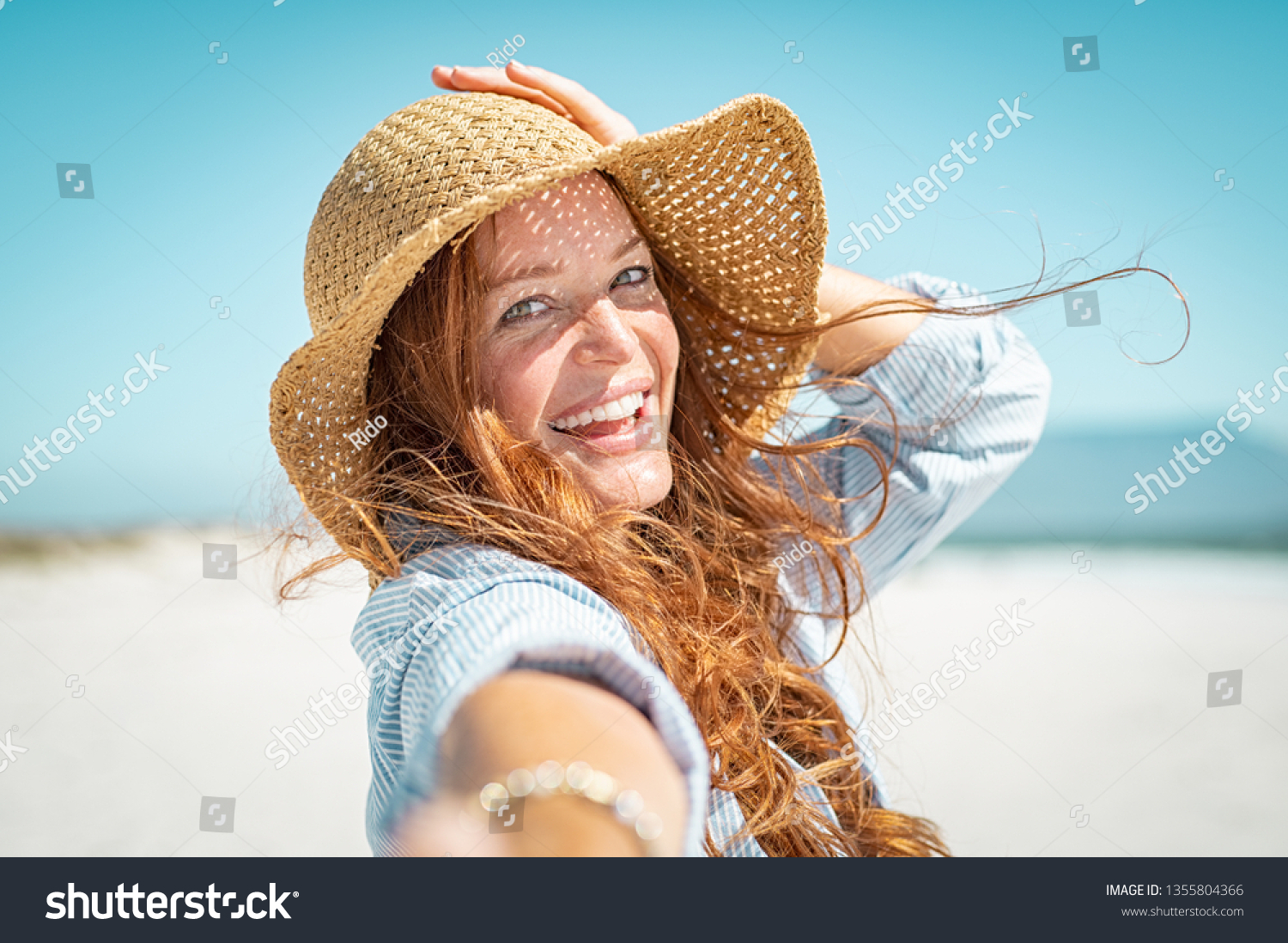 Portrait of beautiful mature woman in casual wearing straw hat at seaside. Cheerful young woman smiling at beach during summer vacation. Happy girl with red hair and freckles enjoying the sun. #1355804366