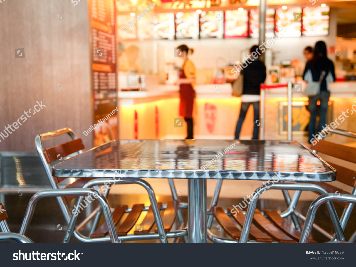 Metal table and chairs at Chinese fastfood cafe in foodcourt with blurred counter menu and tray, focus on furniture - public catering background #1355819039