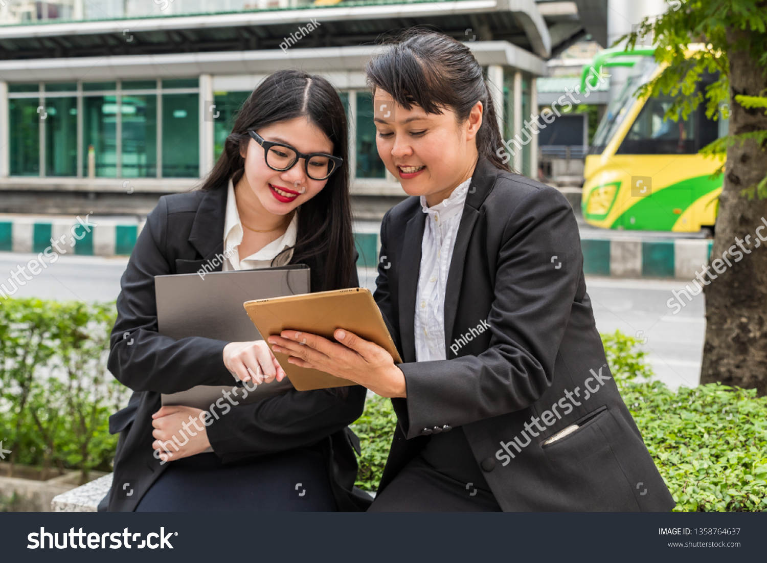 Two businesswomen looking at tablet while holding paper file holder with cityscape as background #1358764637