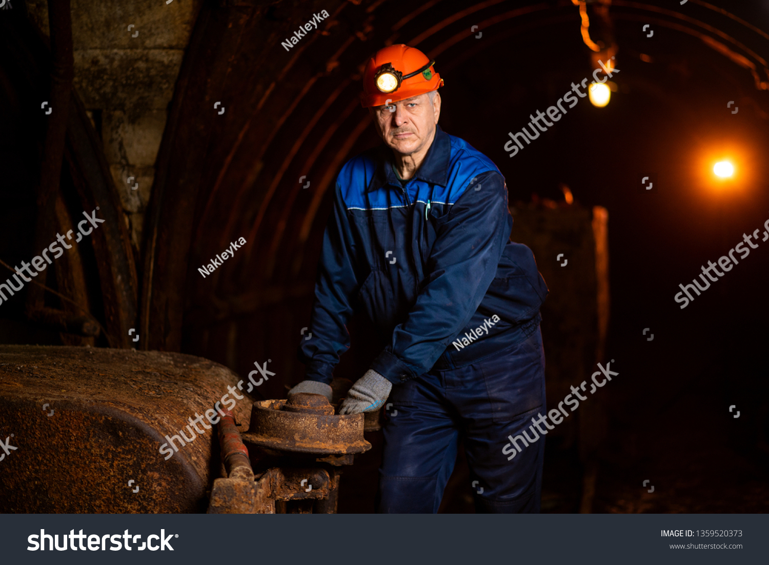 An elderly man dressed in work overalls and a helmet stands near the old inverted vogonetki. Mine worker. Miner #1359520373