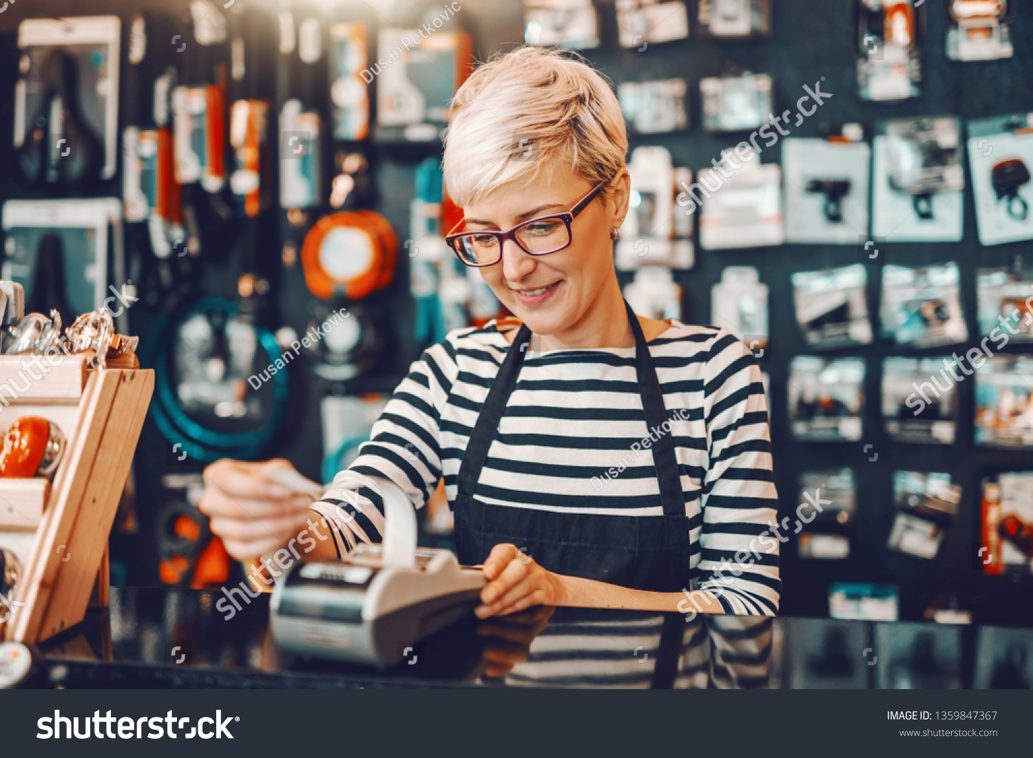Smiling Caucasian female worker with short blonde hair and eyeglasses using cash register while standing in bicycle store. #1359847367