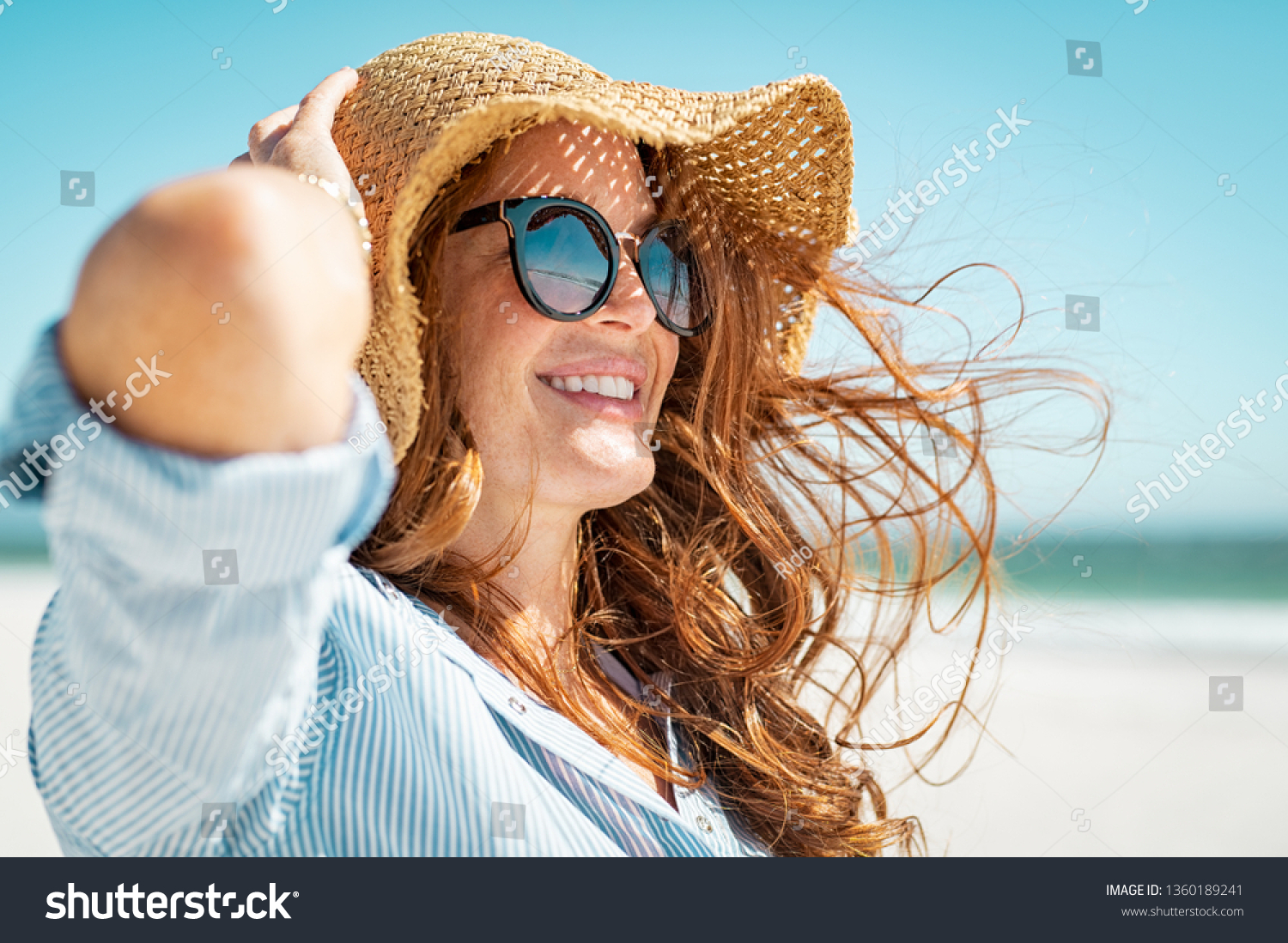 Side view of beautiful mature woman wearing sunglasses at beach. Young smiling woman on vacation looking away while enjoying sea breeze wearing straw hat. Closeup portrait of attractive girl relaxing. #1360189241