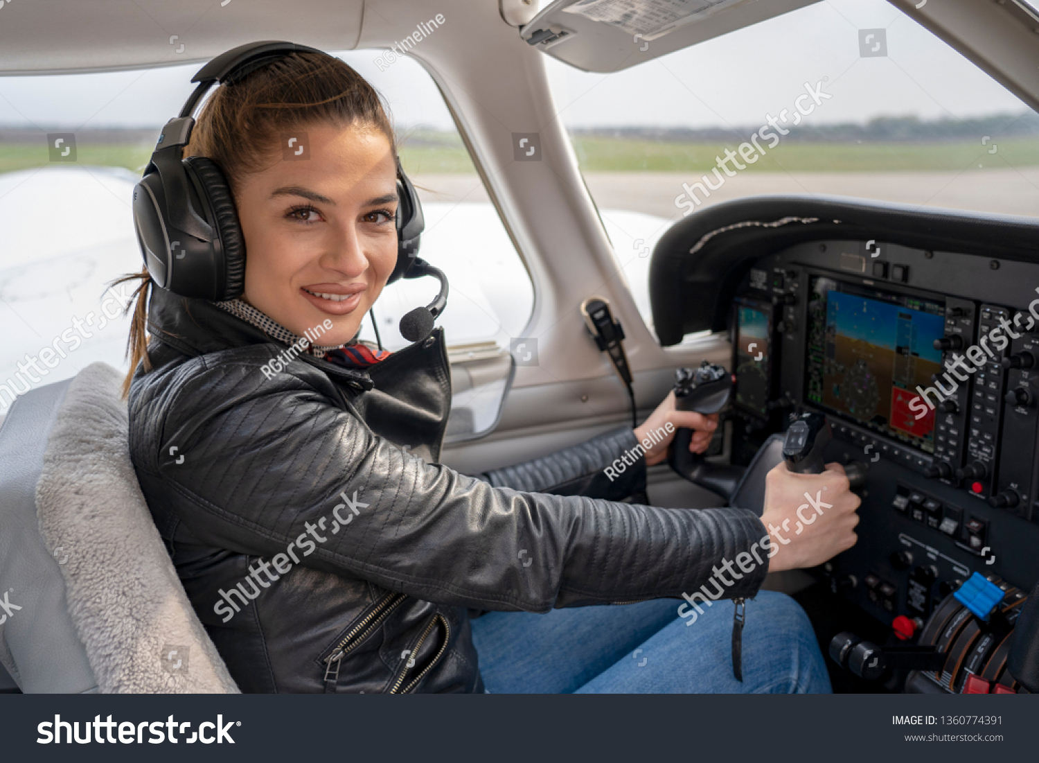 Beautiful Smiling Young Woman Pilot With Headset Sitting in Cabin of Modern Aircraft.She is Looking at Camera and Holding Steering Wheel of a Private Plane. Smiling Female Pilot in the Light Aircraft. #1360774391