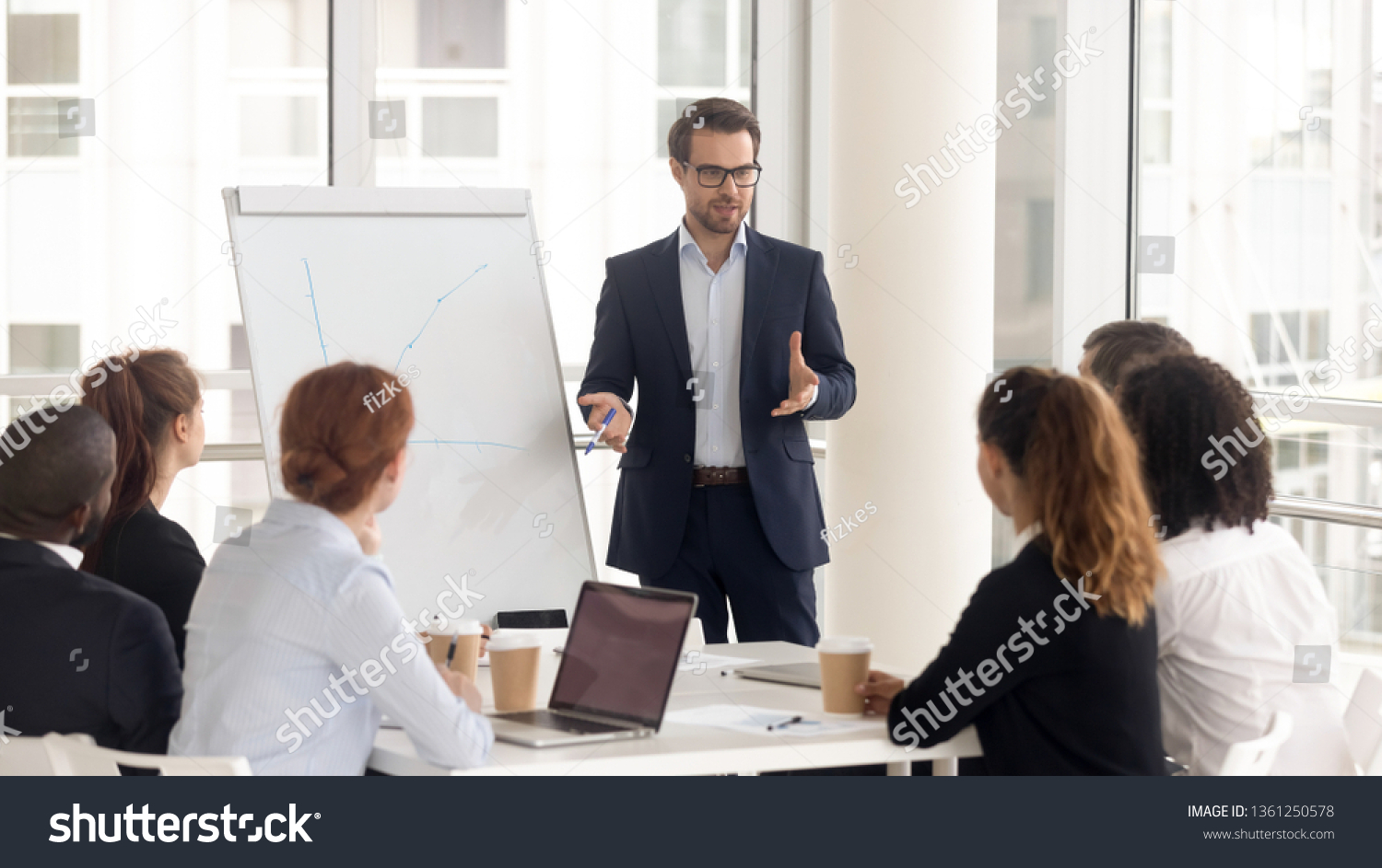 Male business coach speaker in suit give flipchart presentation, speaker presenter consulting training persuading employees client group, mentor leader explain graph strategy at team meeting workshop #1361250578