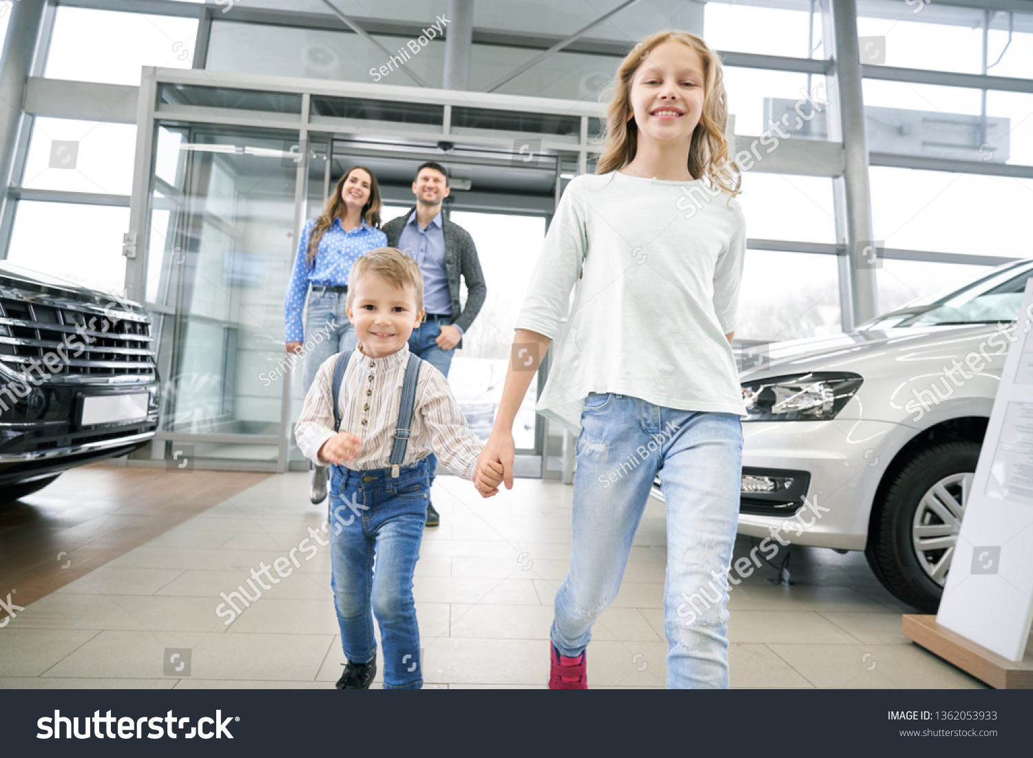 Front view of funny little boy with elder sister entering in big auto showroom. Cute children with parents looking at new cars, choosing and testing automobiles. Concept of car buying. #1362053933