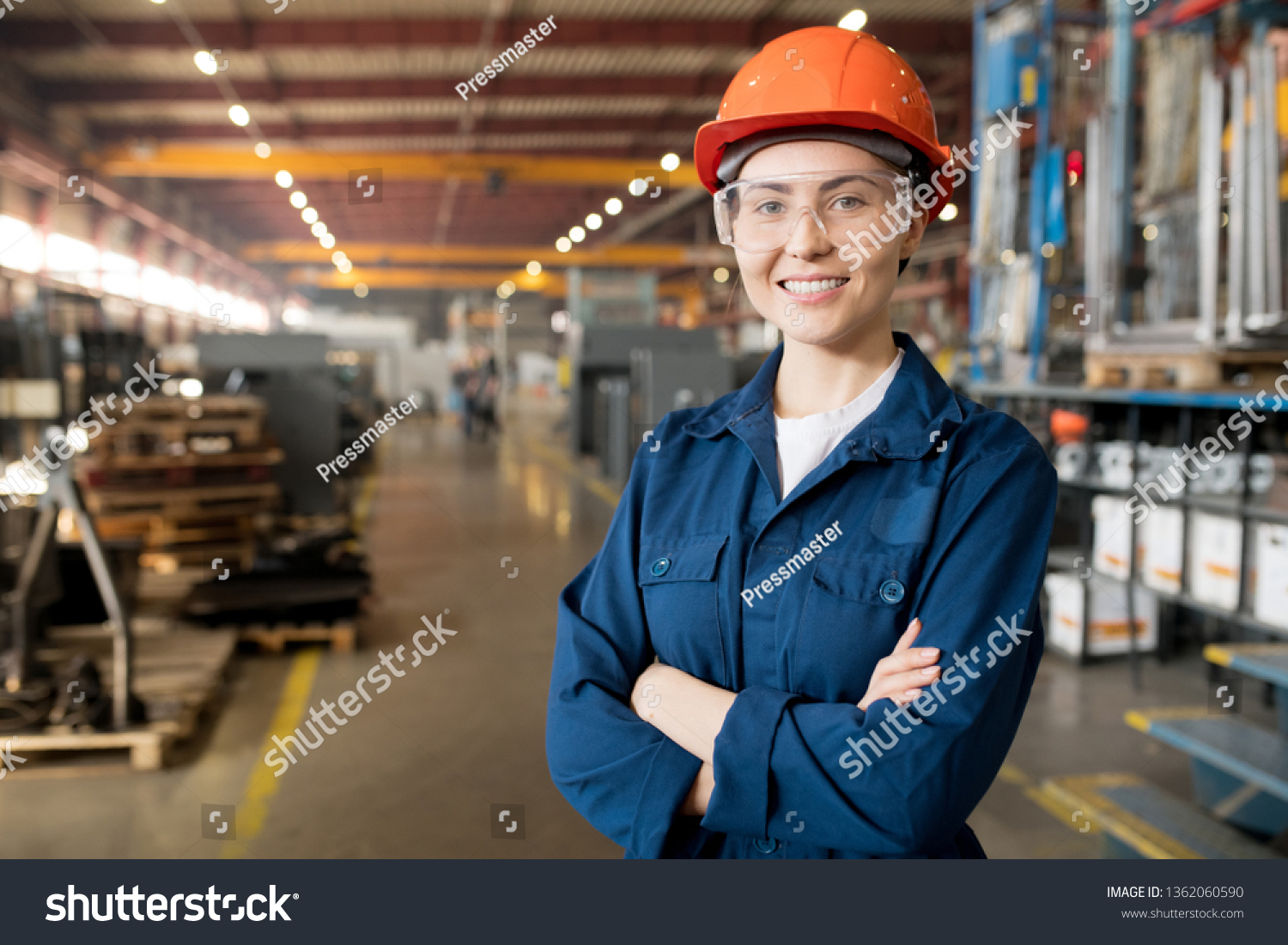Young smiling female technician in blue uniform, protective eyeglasses and helmet working in modern factory #1362060590
