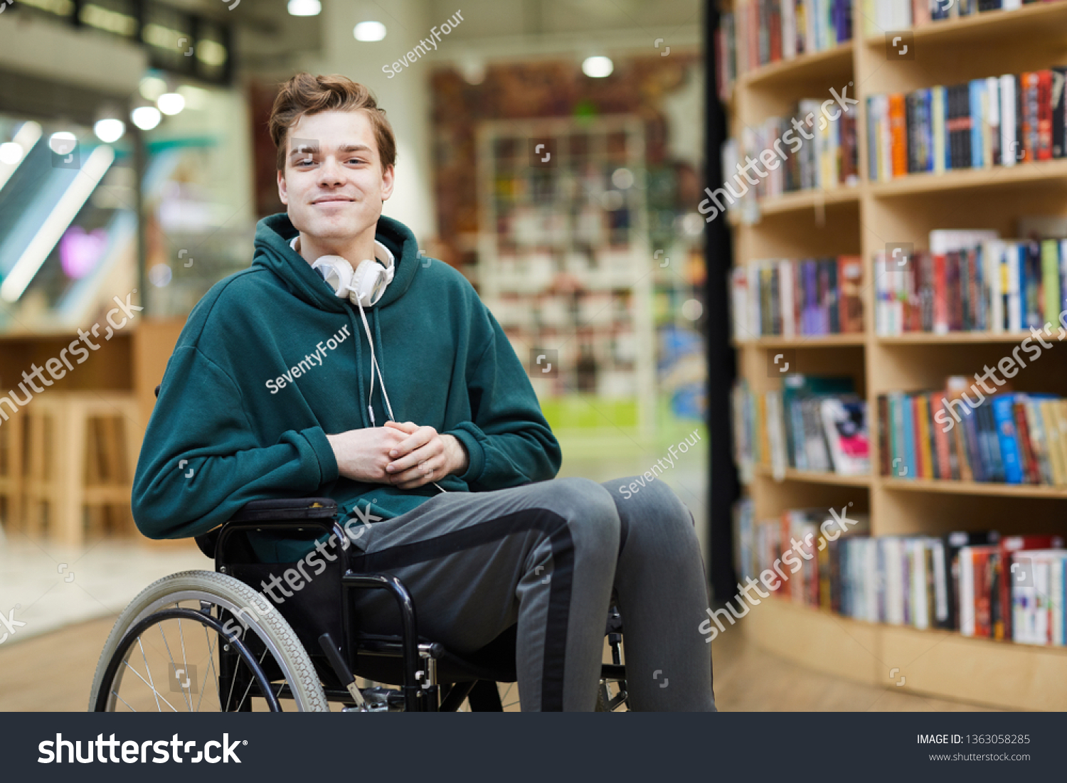 Content handsome young disabled student with headphones on neck siting in wheelchair and looking at camera in modern library or bookstore #1363058285