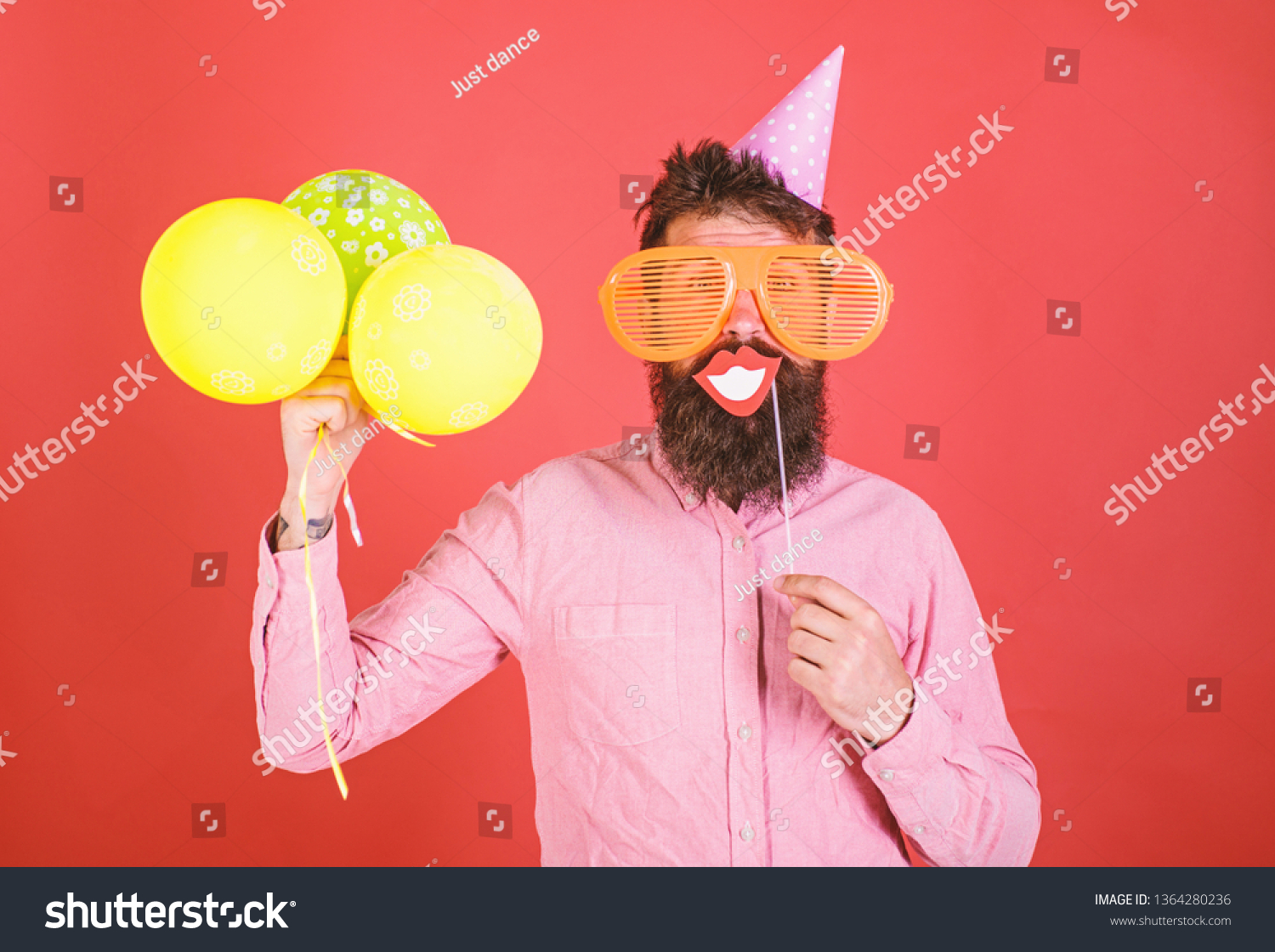 Hipster in giant sunglasses celebrating. Guy in party hat with air balloons celebrates. Man with beard on cheerful face holds smiling mouth on stick, red background. Photo booth fun concept. #1364280236