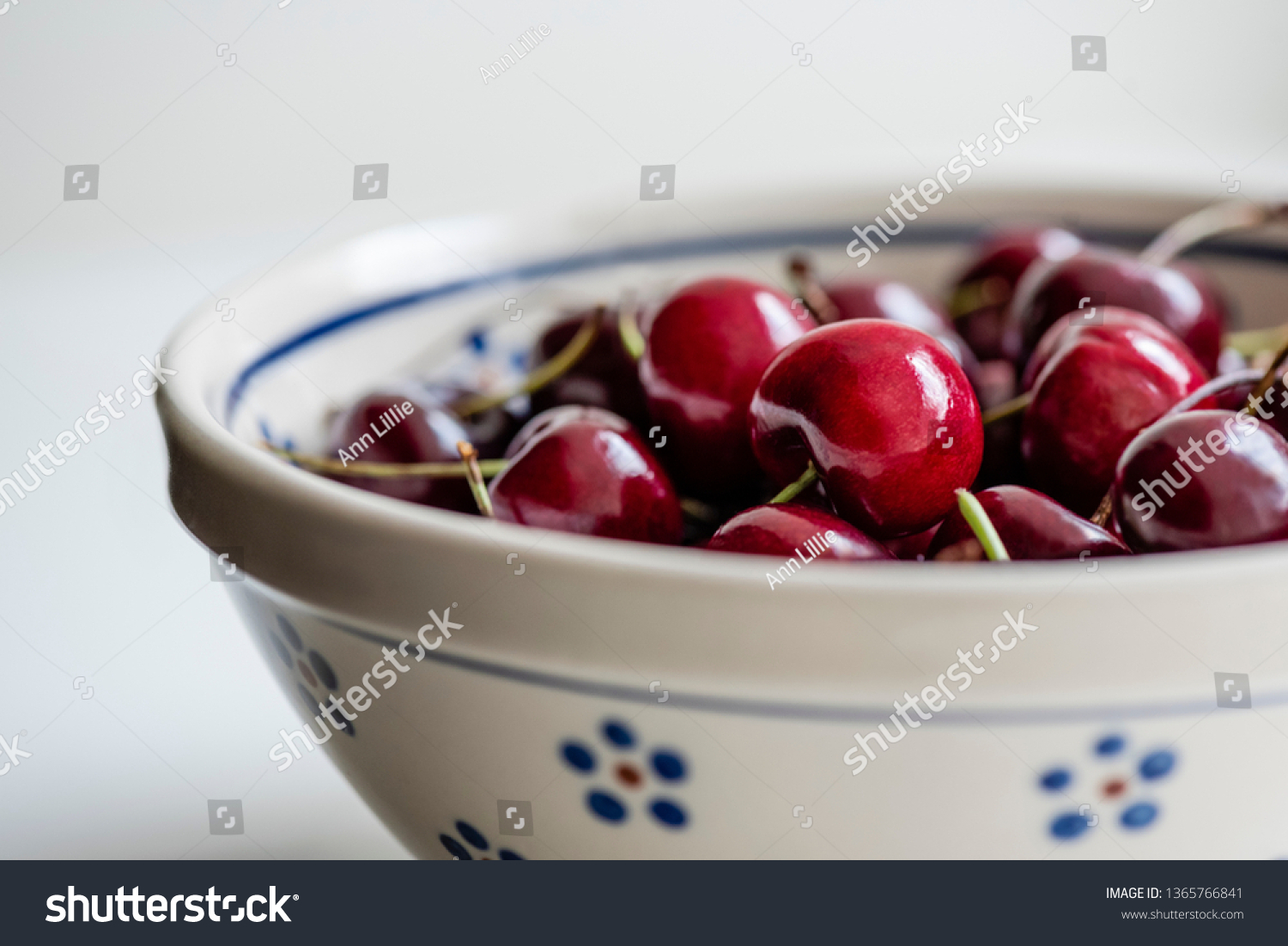Sweet, ripe, dark red bing cherries on stems are grouped in an off-white, round Polish pottery bowl for a morning or afternoon snack. Macro, close-up photo highlighting one shiny cherry. #1365766841