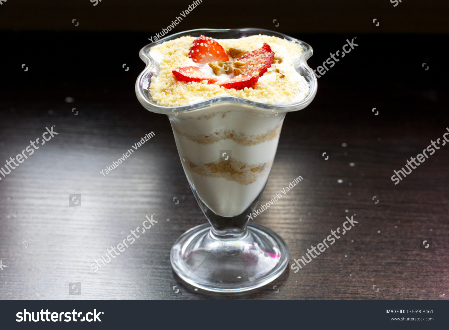 Dessert in a glass goblet. Layers laid biscuit crumbs and cream. Decorated with strawberry slices. With the addition of walnut. On a dark background. #1366908461