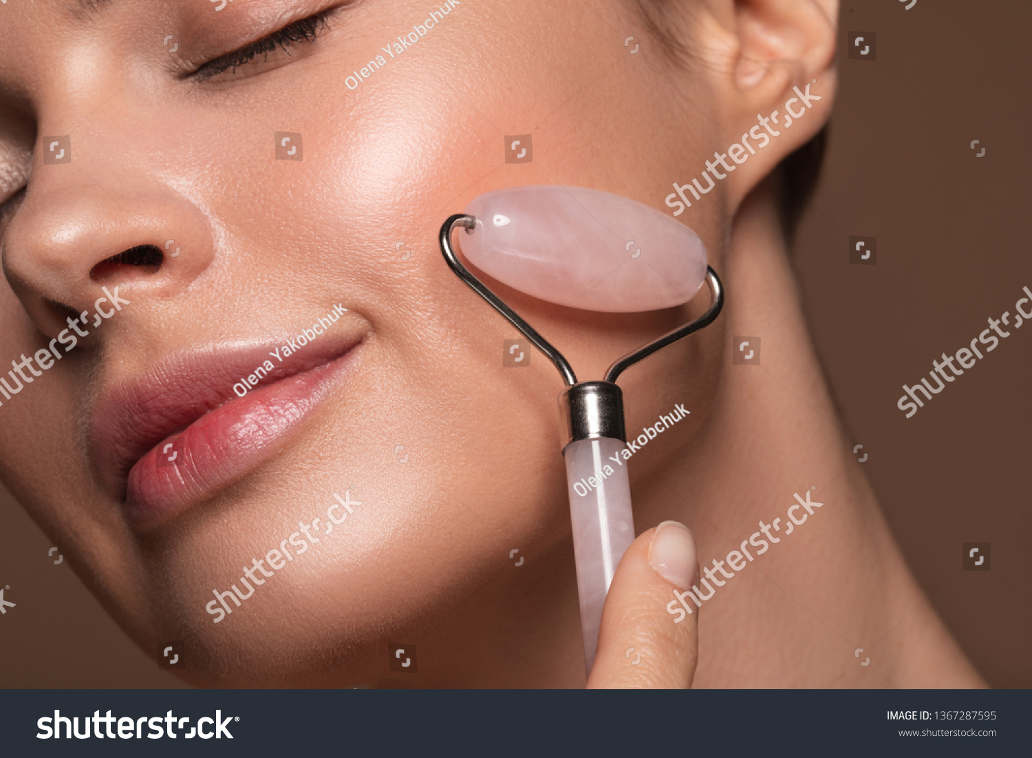 Close up photo of a young woman looking relaxed and smiling while using a natural rose quartz face roller #1367287595