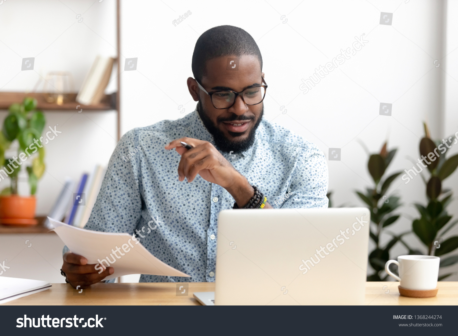 Focused african american businessman working with laptop documents in office holding papers preparing report analyzing work results, black male analyst doing paperwork at workplace using computer #1368244274