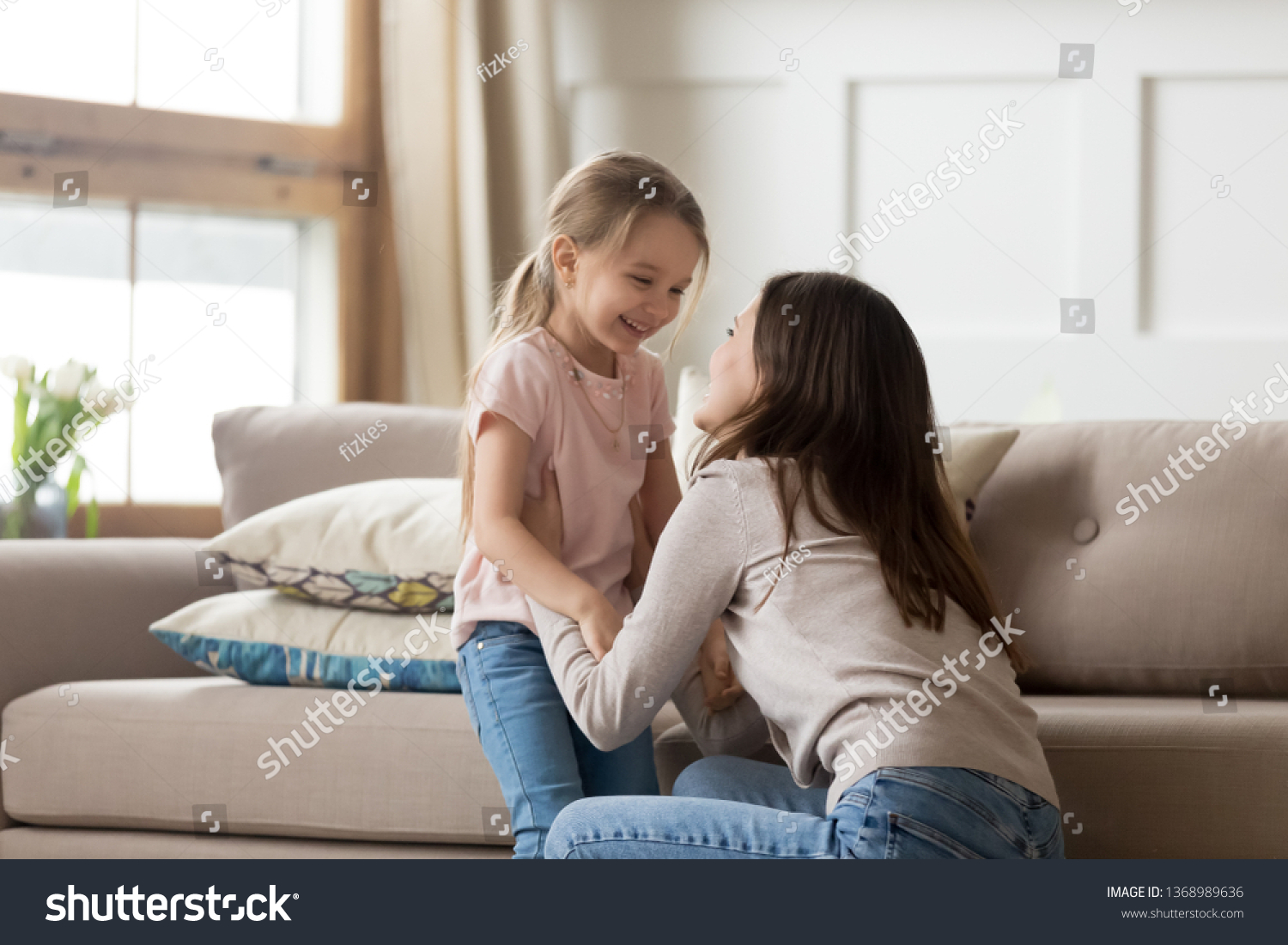 Cheerful daughter looking at overjoyed mother laughing having fun in living room at home, older younger sister enjoy time together spend funny activity on weekend, love and care tender moment concept #1368989636