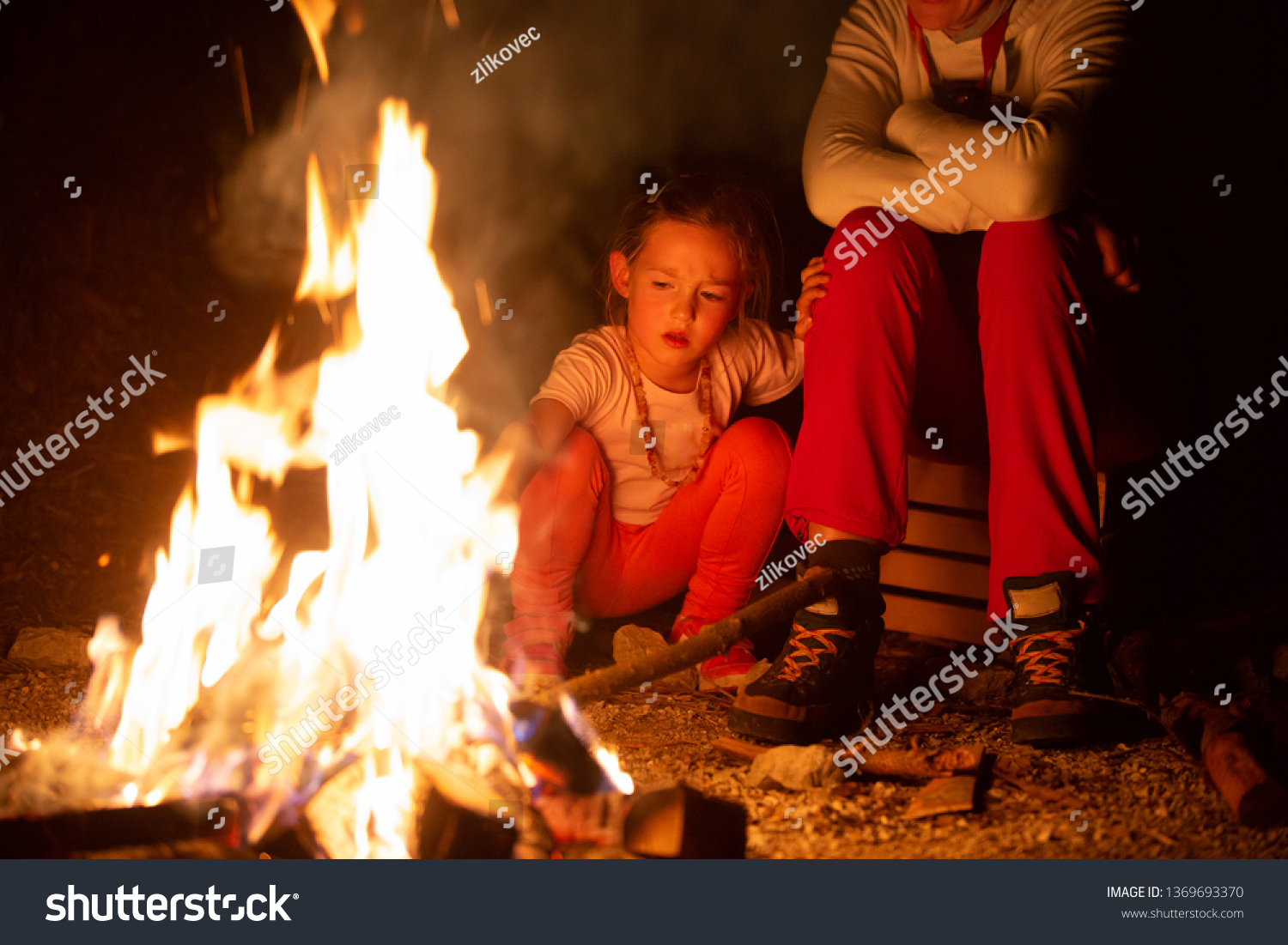Mother and daughter spending quality time by a self-made campfire during adventurous camping trip, playing with fire. Active natural lifestyle, family time and love concept.  #1369693370