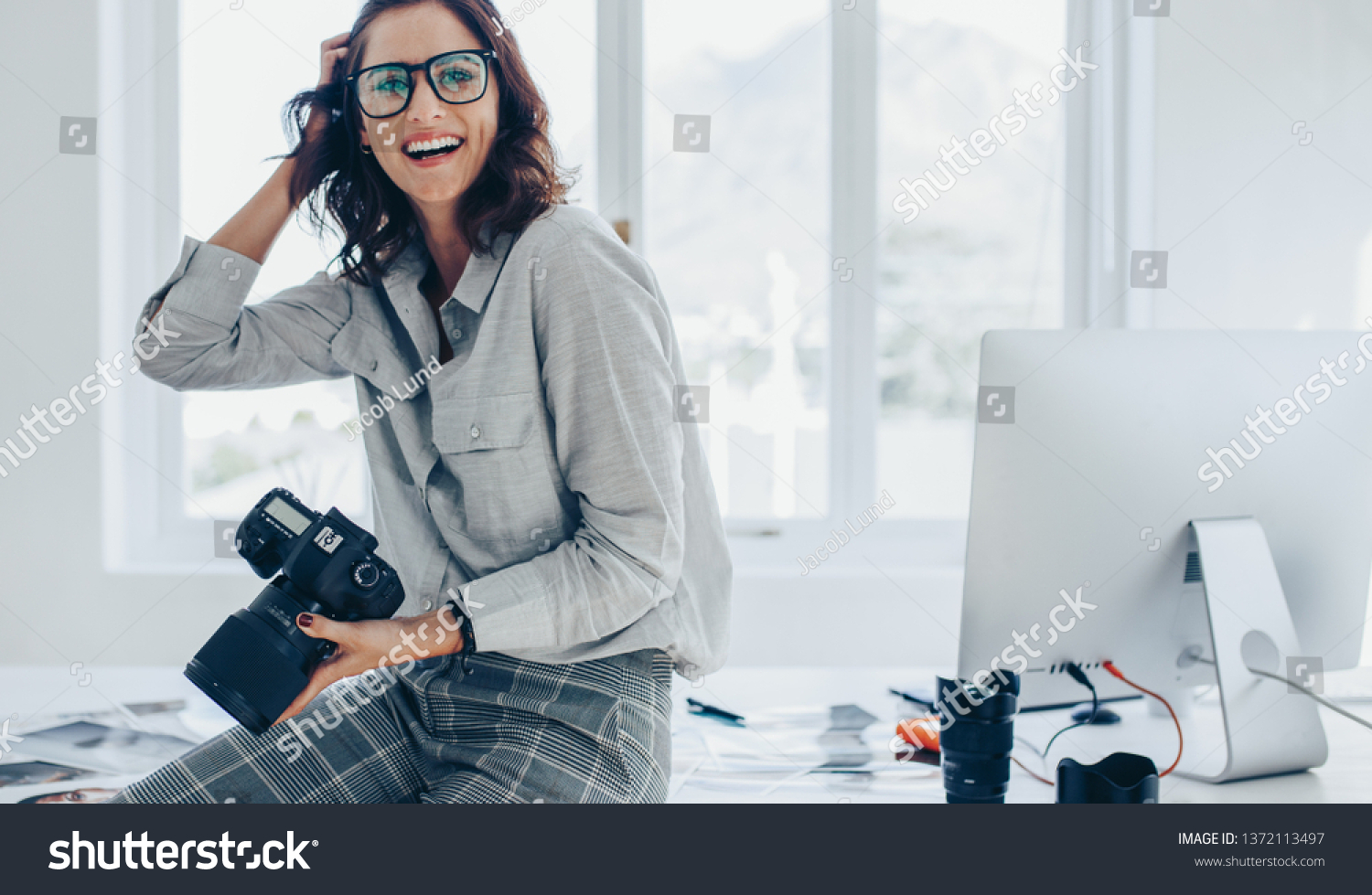 Smiling female photographer with a professional camera sitting on her desk. Woman with dslr camera in office looking away and smiling. #1372113497