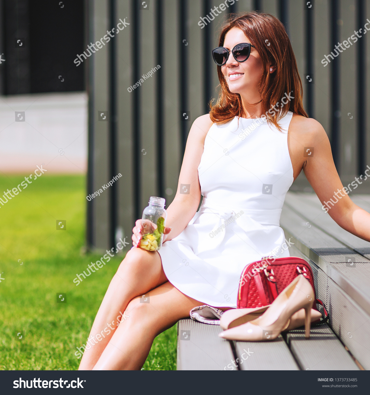 Outdoors lifestyle fashion portrait of pretty red hair girl sitting on the bench. Drinking natural lemonade. Enjoing sunny day, resting. Wearing stylish white dress, sunglass. Shoeless. Rest for feet #1373733485