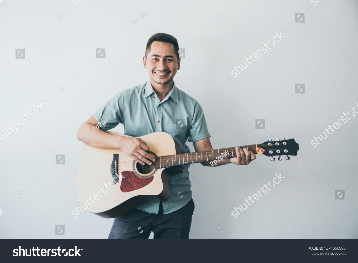 Cheerful guitarist. Cheerful handsome young man playing guitar and smiling while standing on white wall background #1374066995
