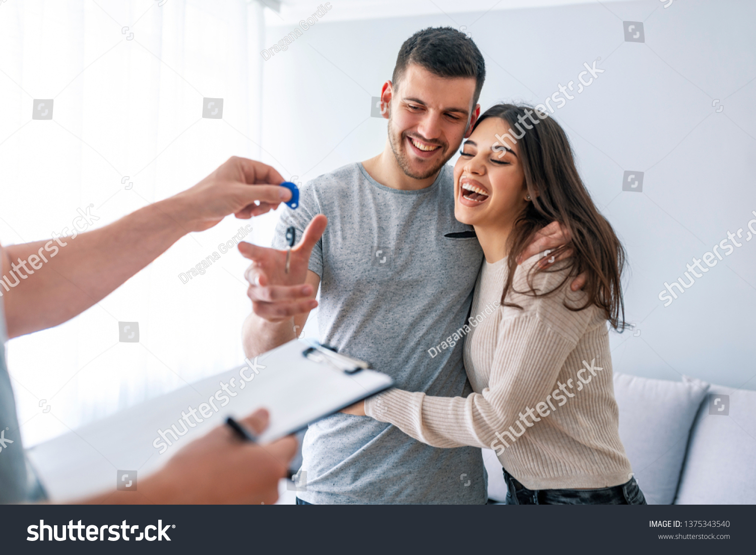 Happy couple is taking keys from their new house from broker and smiling. Hands of estate agent giving keys to the couple. The agent handed the keys a young couple #1375343540