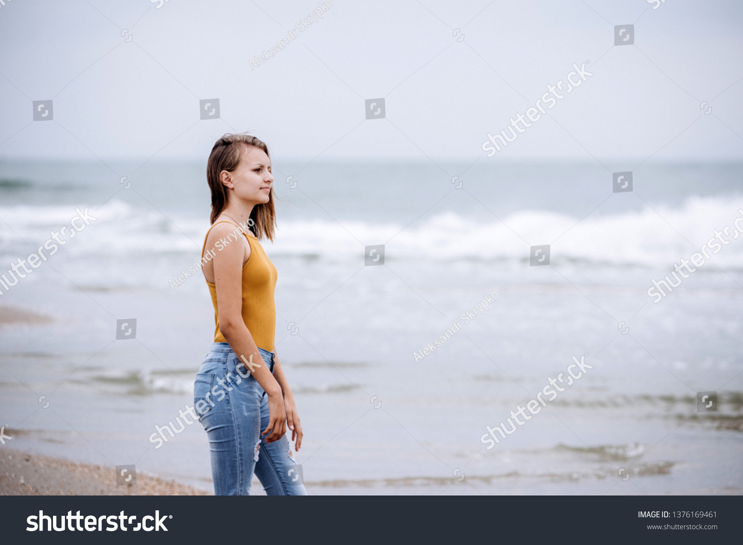 Young beautiful girl walking on the beach on a cloudy morning. She watches as the waves approach the shore.
 #1376169461