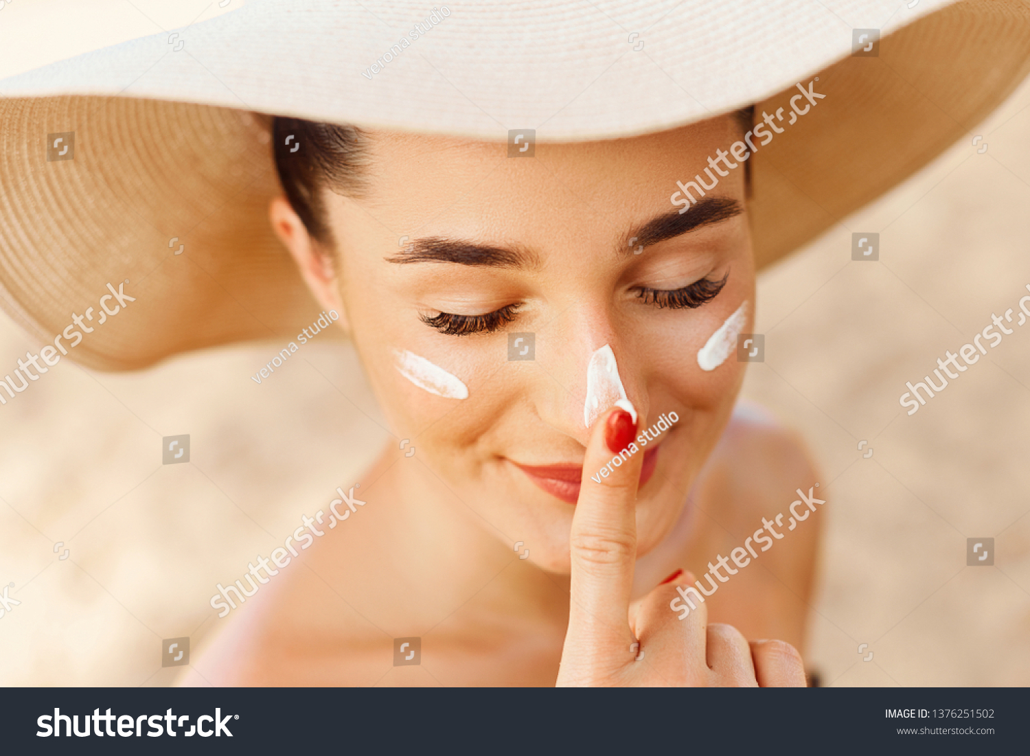 Beautiful Young woman with sun cream on face. Girl holding sunscreen bottle on the beach. Female in hat applying  moisturizing lotion on skin.Skin care. Sun protection. Suntan #1376251502