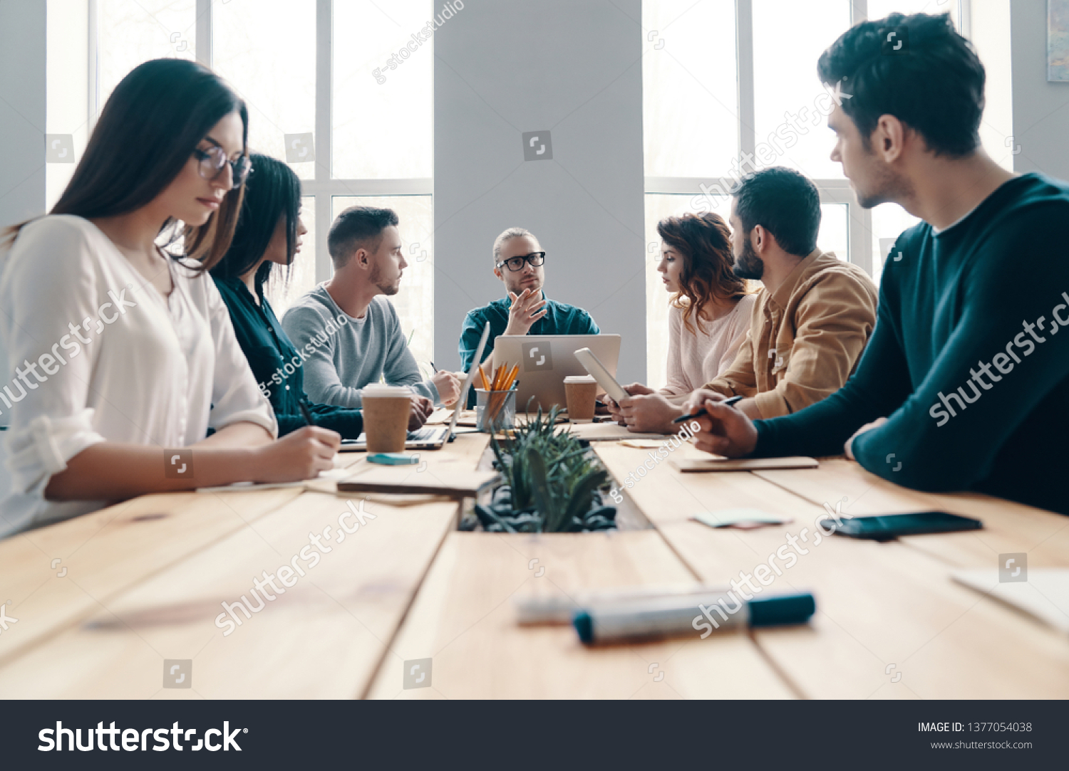 Staff meeting. Group of young modern people in smart casual wear discussing something while working in the creative office           #1377054038
