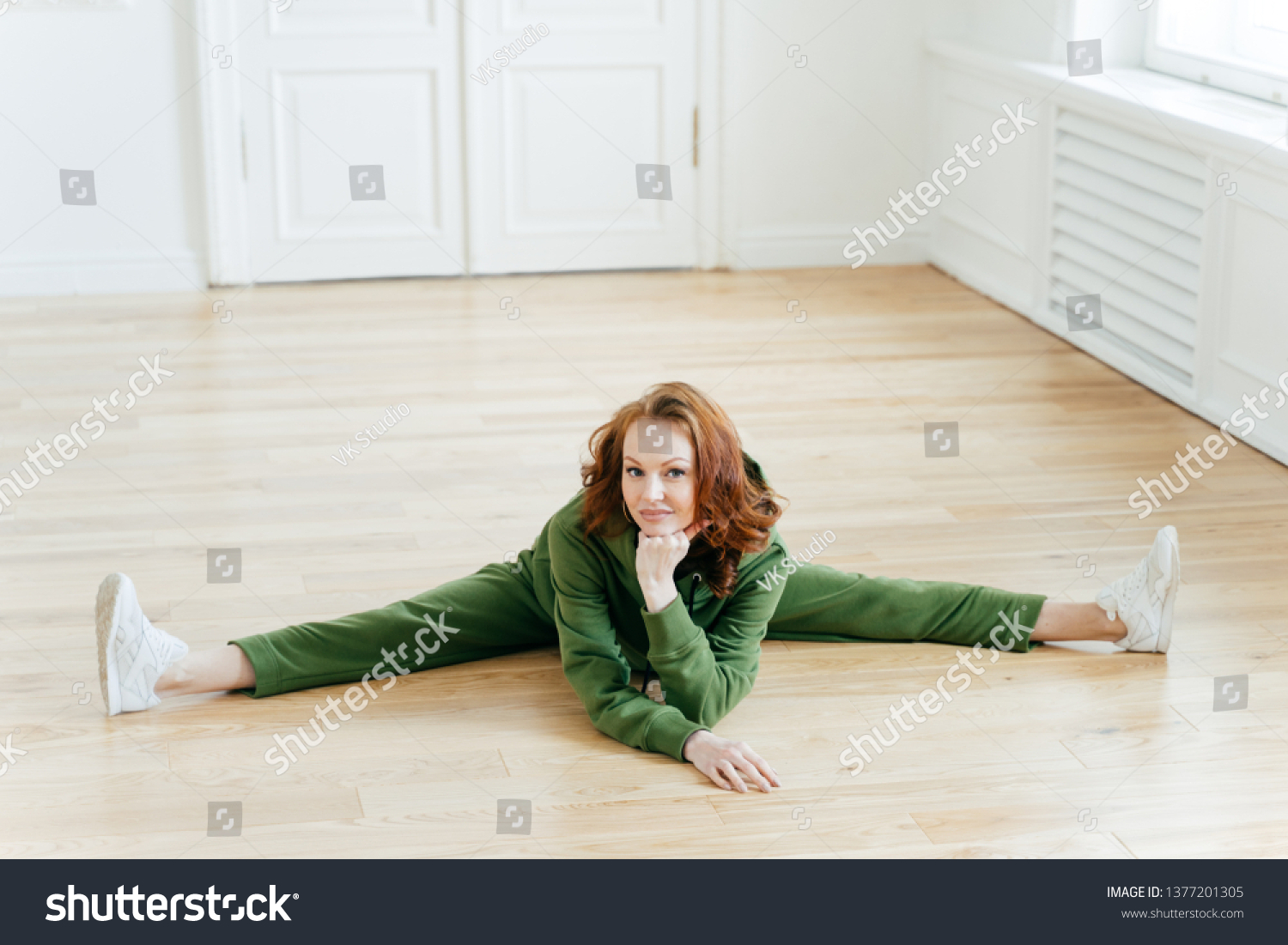 Flexibility, fitness and stretching concept. Attractive redhead female does leg split, stretches legs, wears green sweatsuit, sneakers, keeps hand under chin. Sport gymnast in active wear poses indoor #1377201305