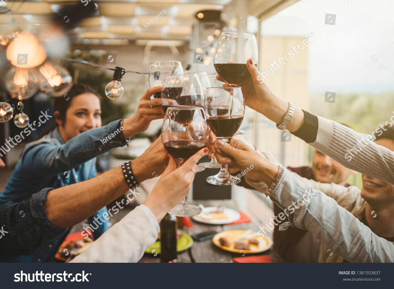 Young friends celebrating at dinner at sunset - Detail of hands while toasting with glasses of wine - Happy people at a terrace party after the harvest before sunset - Concept of friendship #1381553837