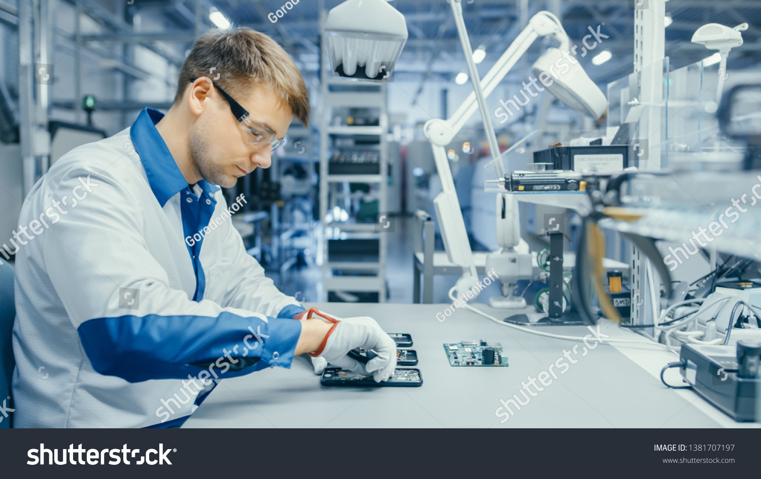 Young Man in Blue and White Work Coat is Using Plier to Assemble Printed Circuit Board for Smartphone. Electronics Factory Workers in a High Tech Factory Facility. #1381707197