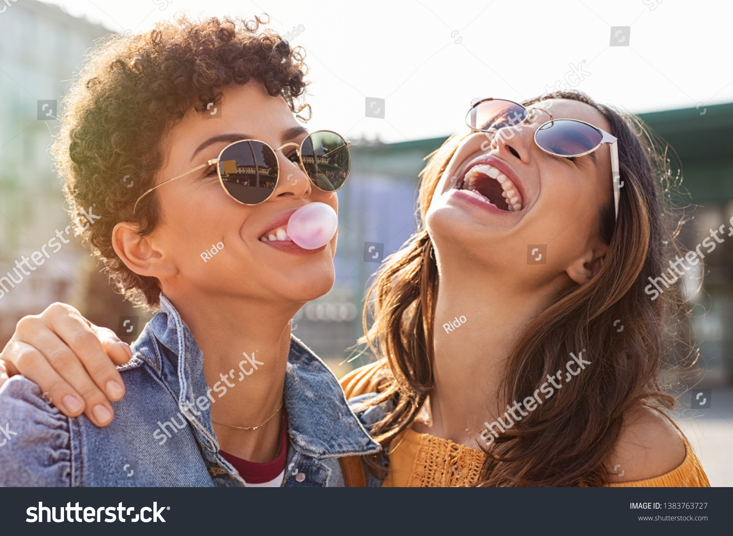 Young latin woman laughing while friend inflating bubble gum. Closeup face of multiethnic friends enjoying outdoor street. Brazilian girl laughing and blowing chewing gum with friend embracing her. #1383763727