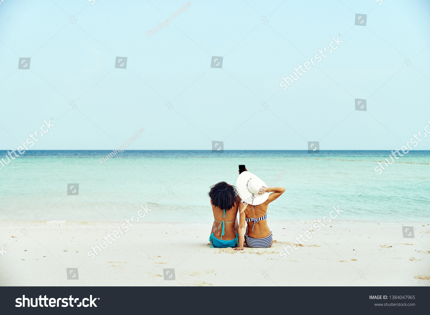Rearview of two young female friends wearing bikinis and taking selfies while suntanning on a sandy beach during a tropical vacation #1384047965