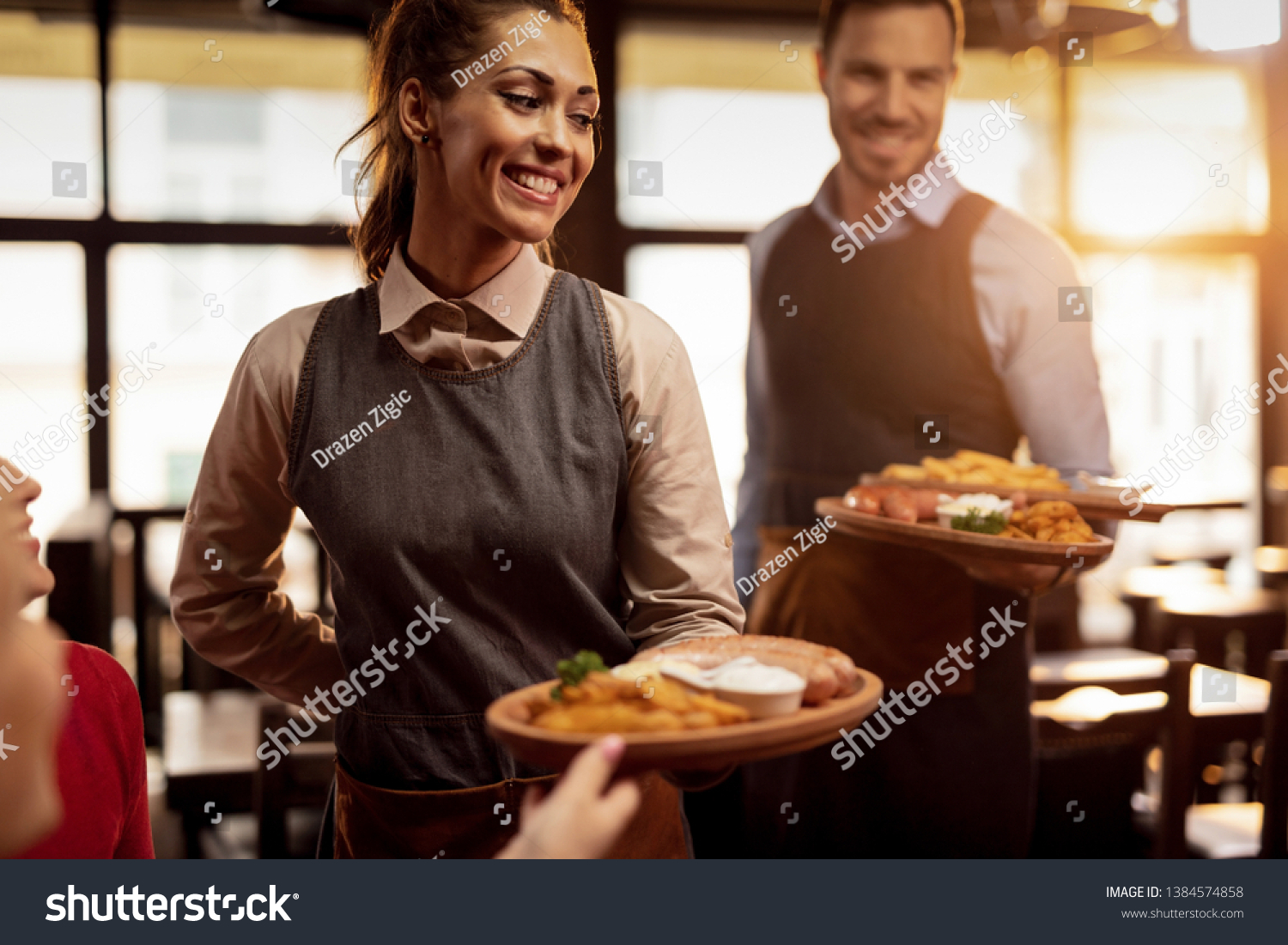 Two waiters serving lunch and brining food to their gusts in a tavern. Focus is on happy waitress.  #1384574858