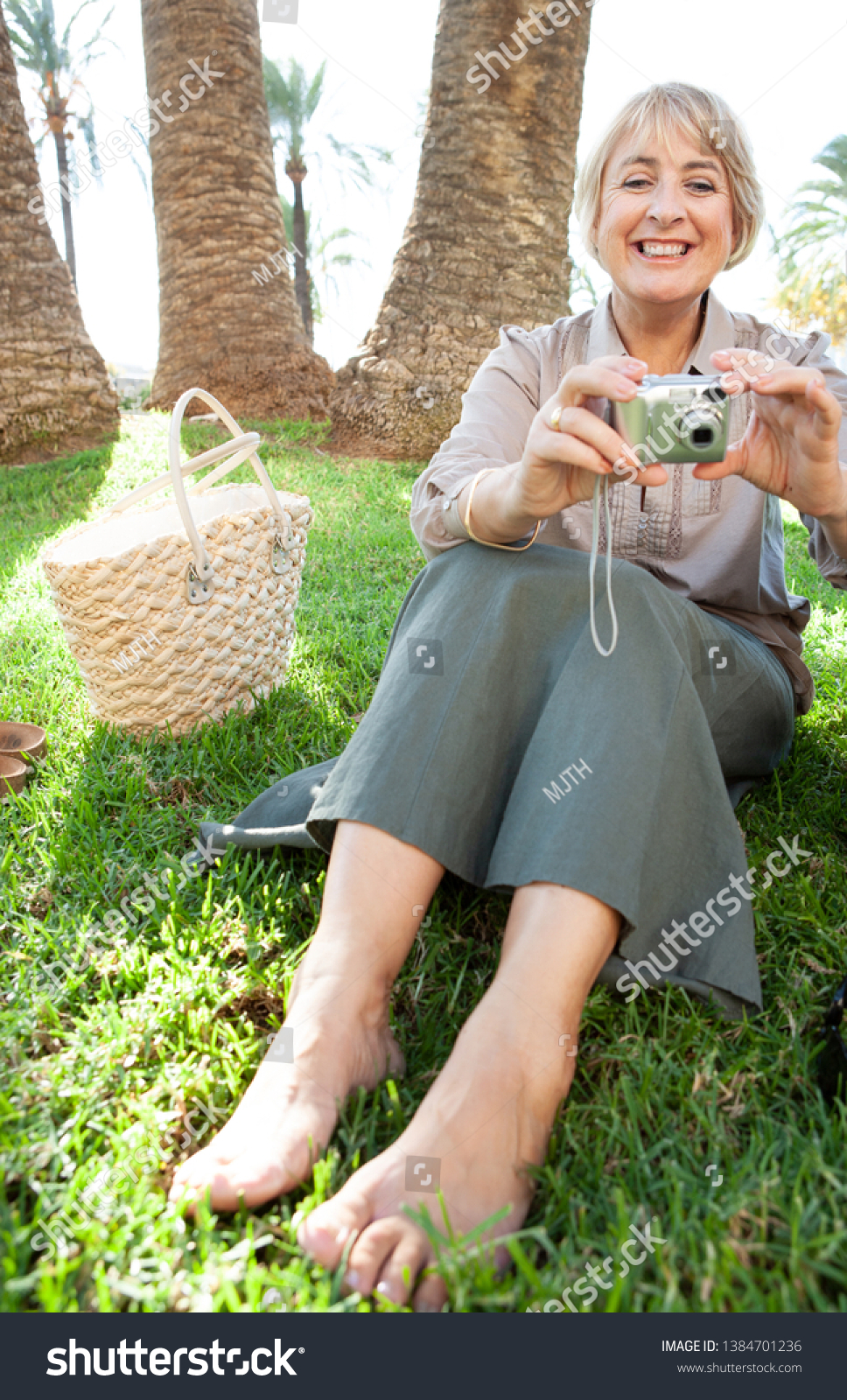 Beautiful mature woman sitting relaxing on grass park holding a digital photographic camera, smiling in sunny outdoors. Senior female tourist on holiday break, leisure technology lifestyle leisure. #1384701236