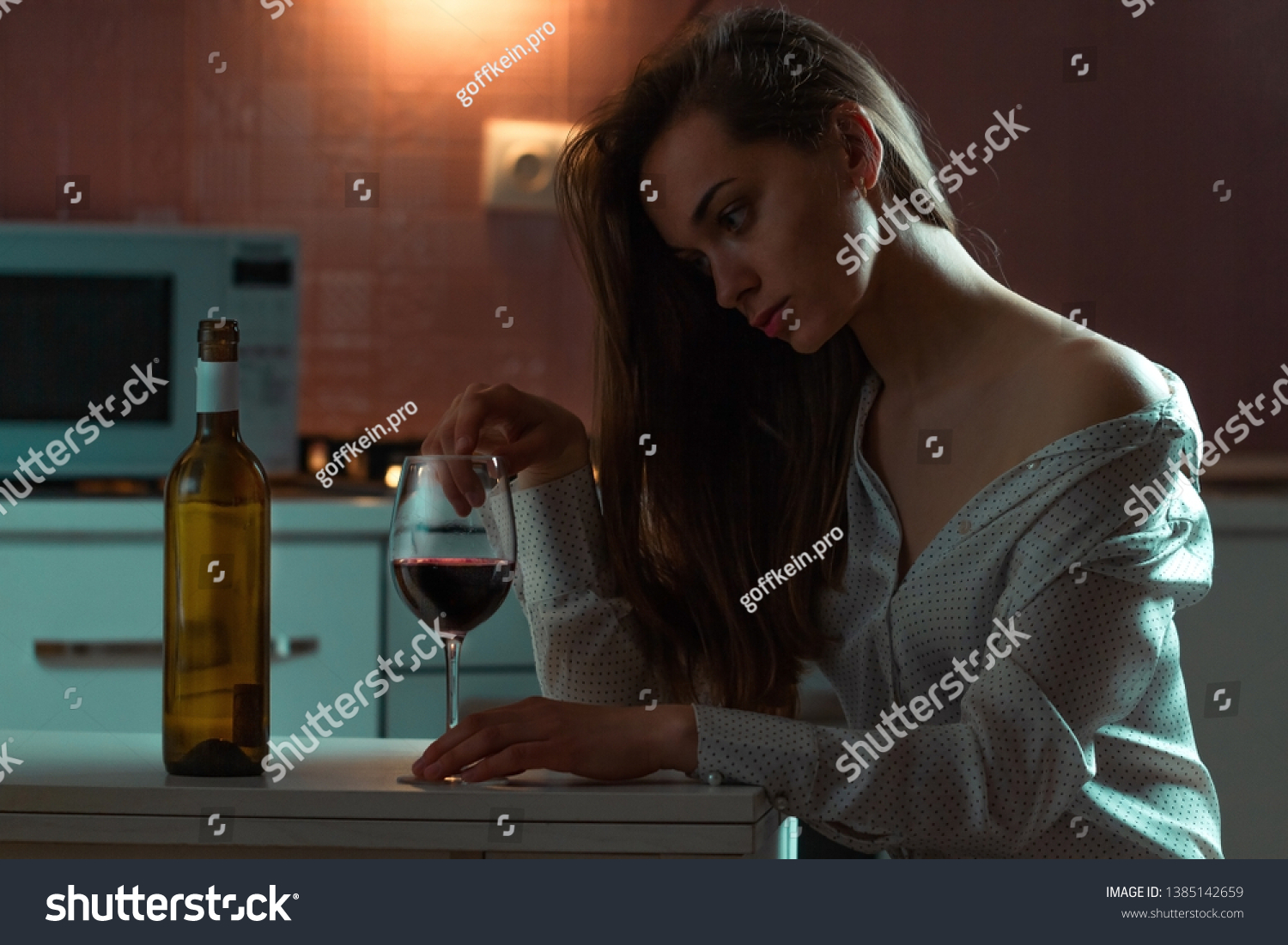 Lonely, sad beautiful young woman in a blouse with alcoholic beverage is drinking alone in evening at home. Female alcoholism and alcohol addiction #1385142659