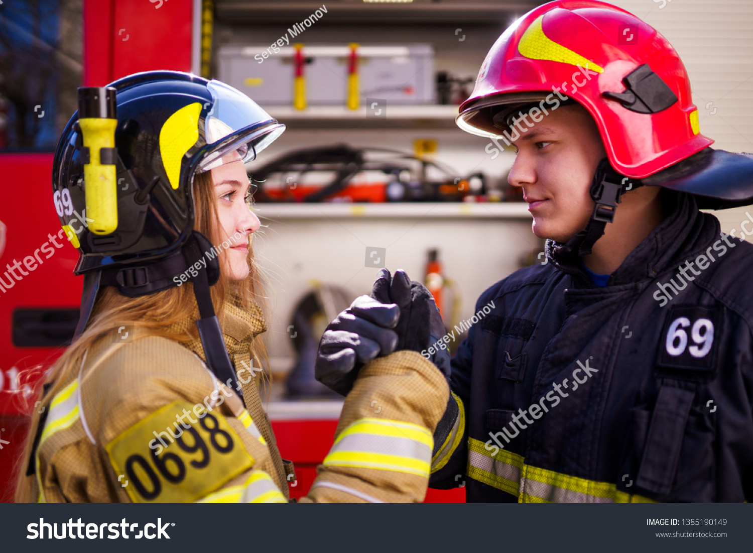 Side view of woman firefighter and man wearing helmets doing a handshake near fire engine #1385190149