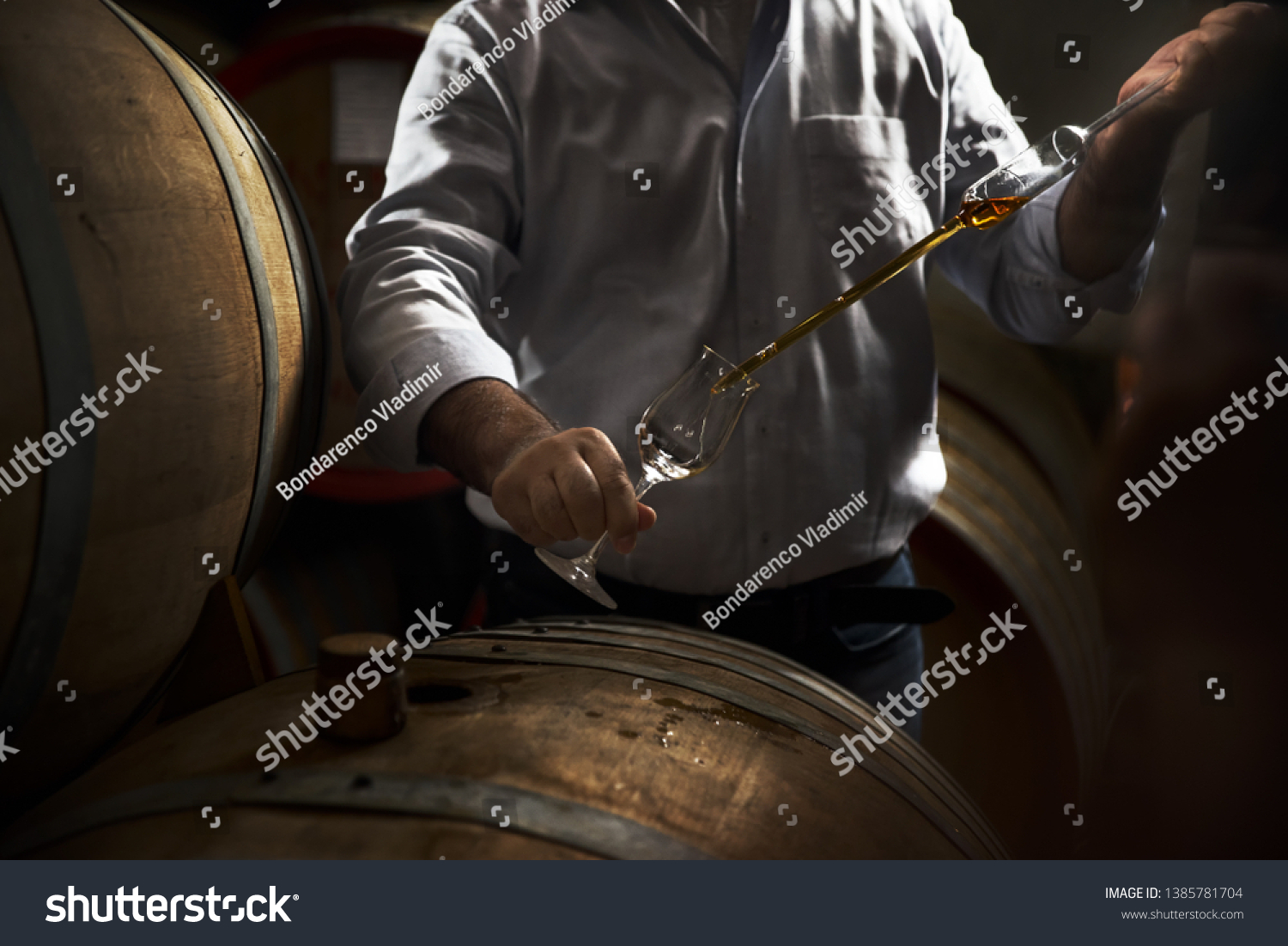 Man, pouring cognac from the barrel into glass in old rustic underground wine cellar with rows of big oak barrels. Famous wine and brandy industrial destination #1385781704
